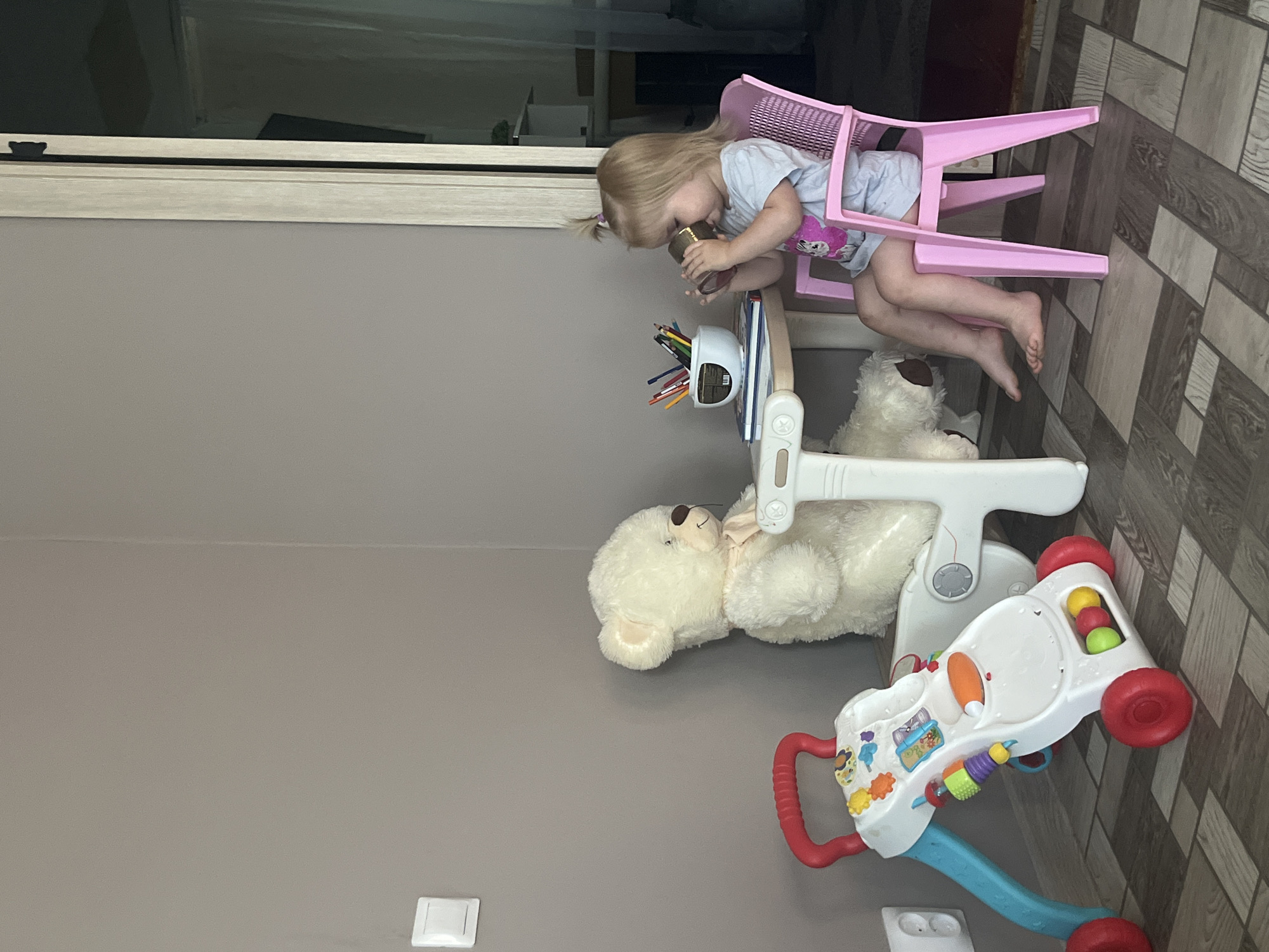 A young child with blonde hair pulled into a ponytail sits on an upside-down pink plastic chair, leaning forward to drink from a small cup. The child wears a light blue t-shirt with a pink design on the front. A toy desk with colored pencils in a dispenser is positioned in front of the child. To the right of the desk, a large, fluffy white teddy bear is lying down. In the foreground, a white and colorful baby walker with red wheels and handles is visible. The scene appears to be indoors, with a grey wall on the left and a tiled floor on the right. A light fixture is visible on the grey wall. The image is oriented vertically.