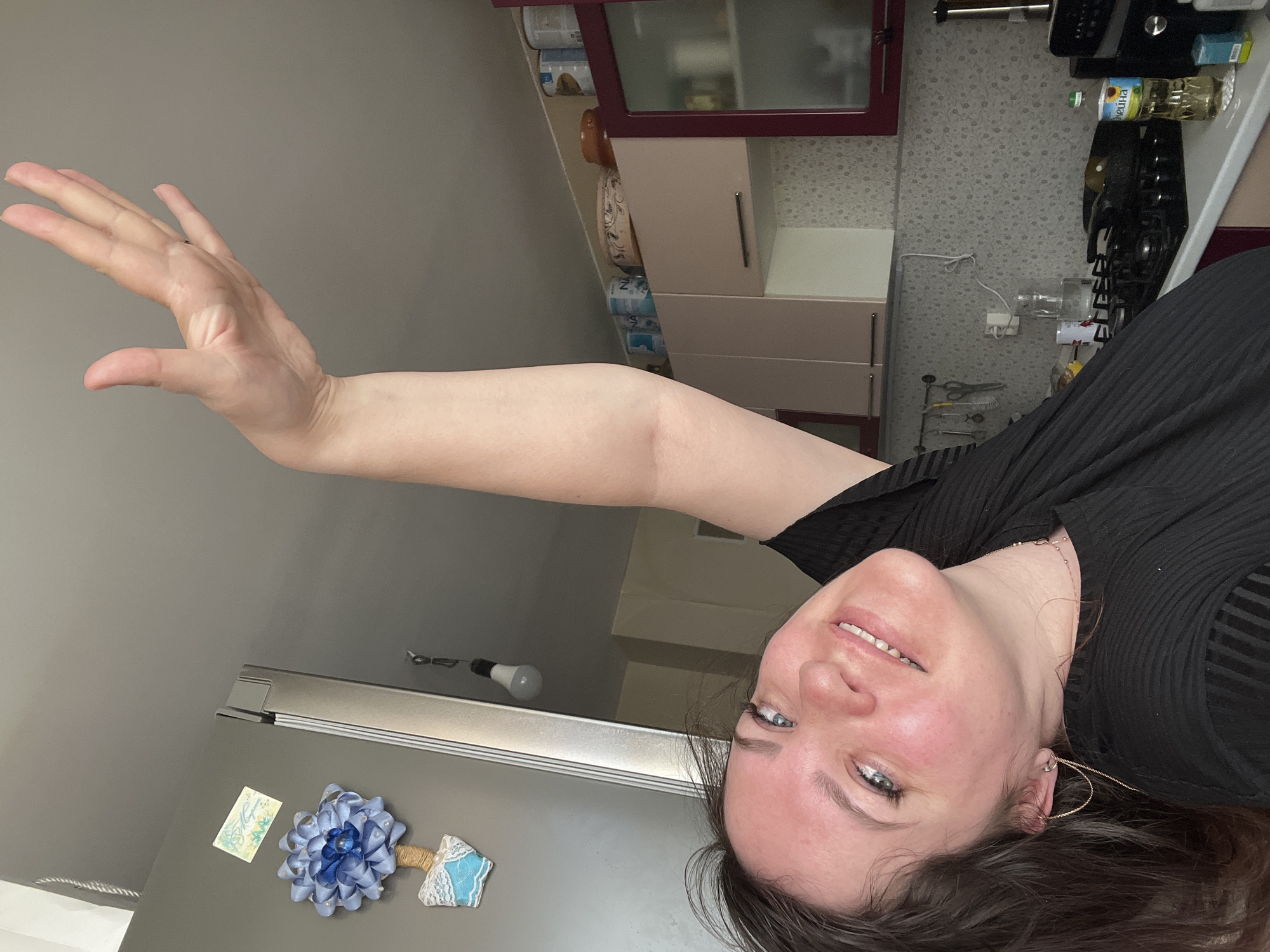 A woman with light skin, blue eyes, and dark hair is smiling at the camera. She is wearing a black ribbed top and gold hoop earrings. Her right arm is raised with her hand extended, palm facing forward. The background shows a kitchen setting. There are cabinets, a countertop with kitchen appliances and supplies, and a refrigerator with decorative magnets on it. Based on the style of the kitchen and the presence of items with Russian writing (like the sunflower oil), it is possible the location is Russia. There is insufficient information to confirm the city of Noviy.