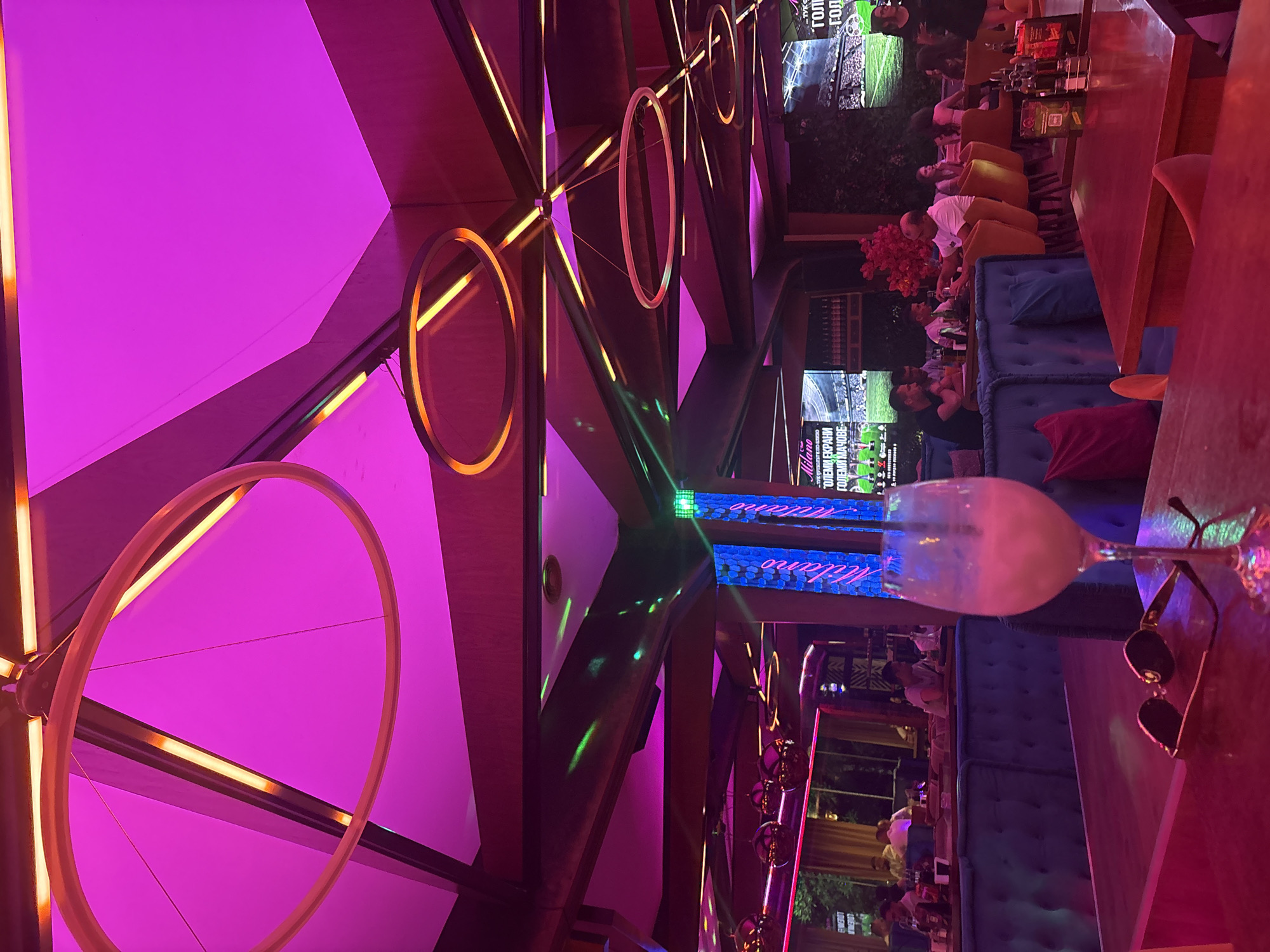 The image shows the interior of a dimly lit restaurant or bar. The ceiling is adorned with geometric panels and illuminated by vibrant pink and orange lights. Several circular light fixtures hang from the ceiling, some with orange accents. The walls are dark, and a few screens are visible in the background, displaying what appears to be a soccer match. The foreground features a table with a drink in a wine glass and a pair of sunglasses. In the midground, patrons are seated at tables and booths, some engaged in conversation, while others are looking towards the screens. The seating is upholstered in blue and orange. The overall atmosphere is modern and somewhat atmospheric, with a focus on colorful lighting.