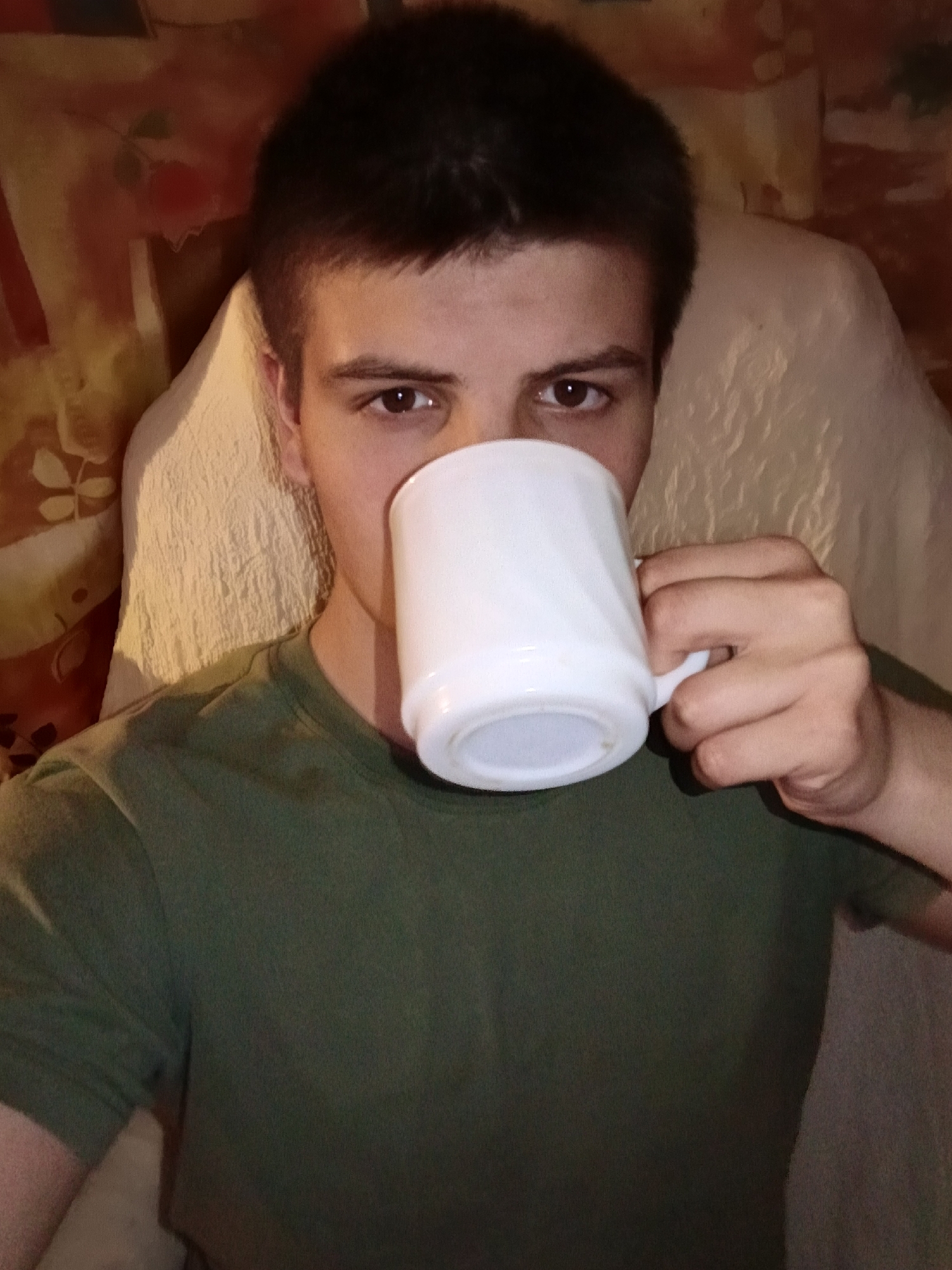 A young man with short dark hair and brown eyes is drinking from a white ceramic mug. He is wearing a green t-shirt. The background includes a pillow or cushion with textured fabric and a patterned surface. The specific location within Красный Маяк, Russia, cannot be determined from the image.