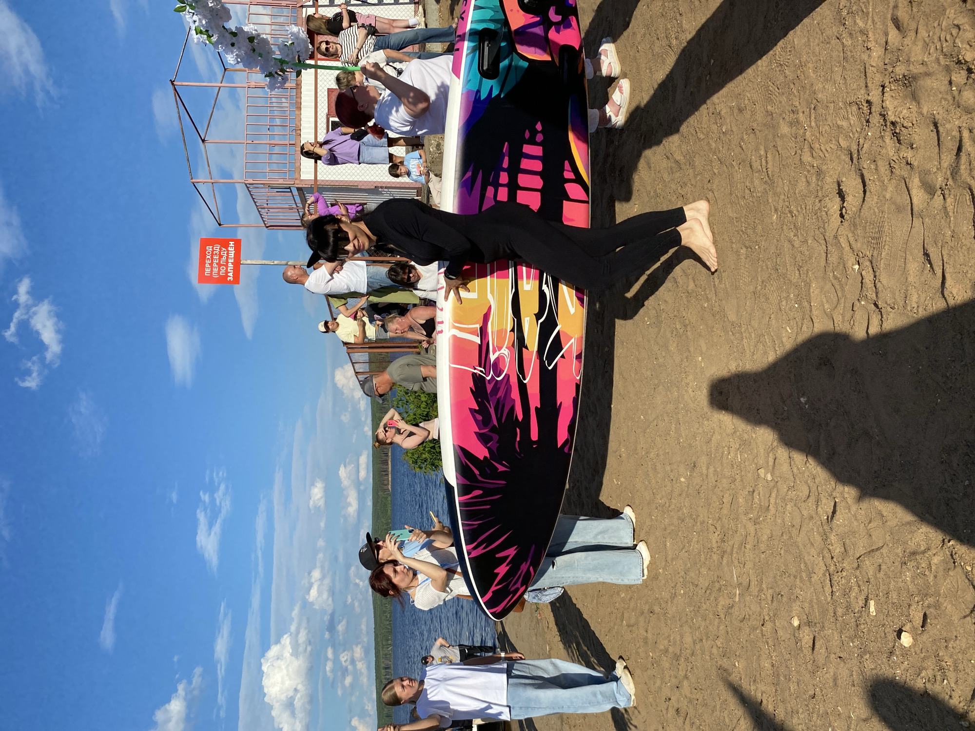 The image depicts a sunny outdoor scene with a group of people on a sandy area near a body of water, likely a lake or river. A bright blue sky with scattered white clouds dominates the upper portion of the image. In the background, a structure that appears to be a viewing platform or observation deck is visible, with a red sign on a pole that reads "ПЕРЕХОД ПО ЛЬДУ ЗАПРЕЩЕН," translating to "Crossing on ice is prohibited." This sign suggests the location might be a recreational area that is used during warmer months.
The main focus of the image is a woman in a black full-body suit holding a colorful surfboard. She is barefoot and positioned in the foreground. To her left, a young woman in light blue jeans and a white t-shirt is also barefoot and holding a surfboard. Another woman in blue jeans and a striped top stands near her, holding a smartphone. Other individuals are scattered in the background, some holding phones and appearing to take pictures. The overall atmosphere is casual and suggests an event or gathering related to water sports or a beach outing. The sandy terrain, the presence of a large body of water, and the people's attire point to a summer setting. The specific location in Chaikovsky, Russia, is inferred from the context of the sign, suggesting this is a public area where such activities are permitted, likely a beach or embankment along the Kama River, which flows through Chaikovsky.