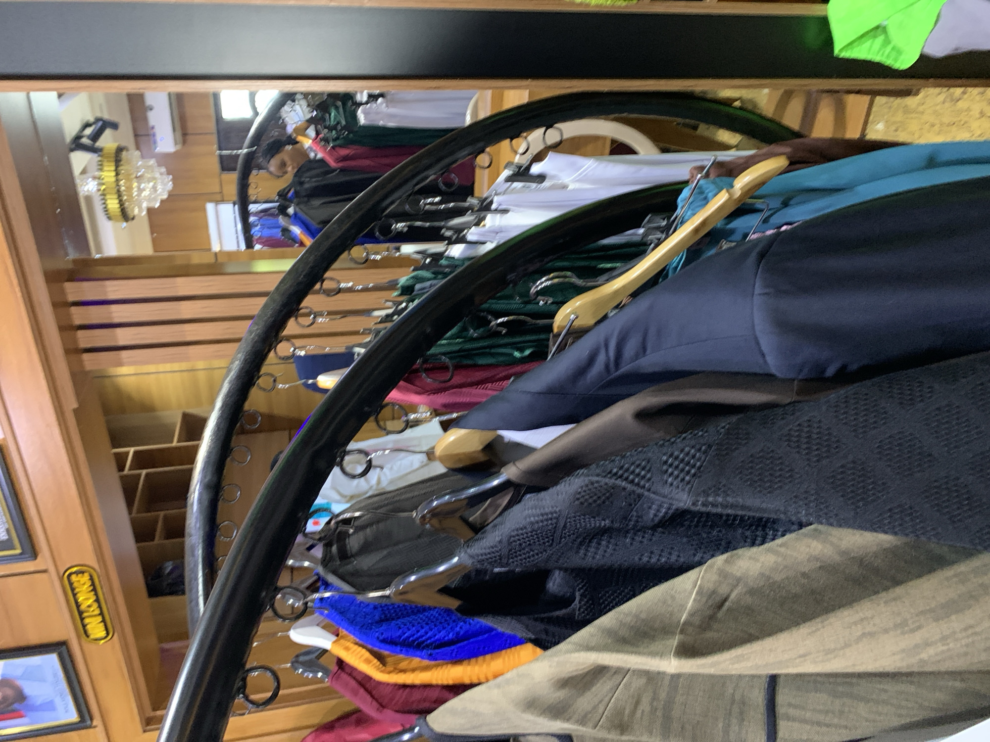 The image depicts a section of a clothing store, likely a boutique, with garments displayed on hangers. The scene appears to be an interior shot, focusing on a rack of men's wear. The clothing includes suits, blazers, and shirts in various colors such as brown, black, blue, and yellow. Some garments are made of textured fabric. The store's interior features wooden shelving units and a curved black metal rack holding the clothes. A mirror in the background reflects a chandelier and part of the store's ceiling, suggesting an interior location. A woman in the background is partially visible, looking towards the clothing. A sign with the text "MINI LOUNGE" is visible on the wall. The overall impression is of a retail space offering formal or semi-formal attire. This scene is situated in Gura Top, Nigeria.