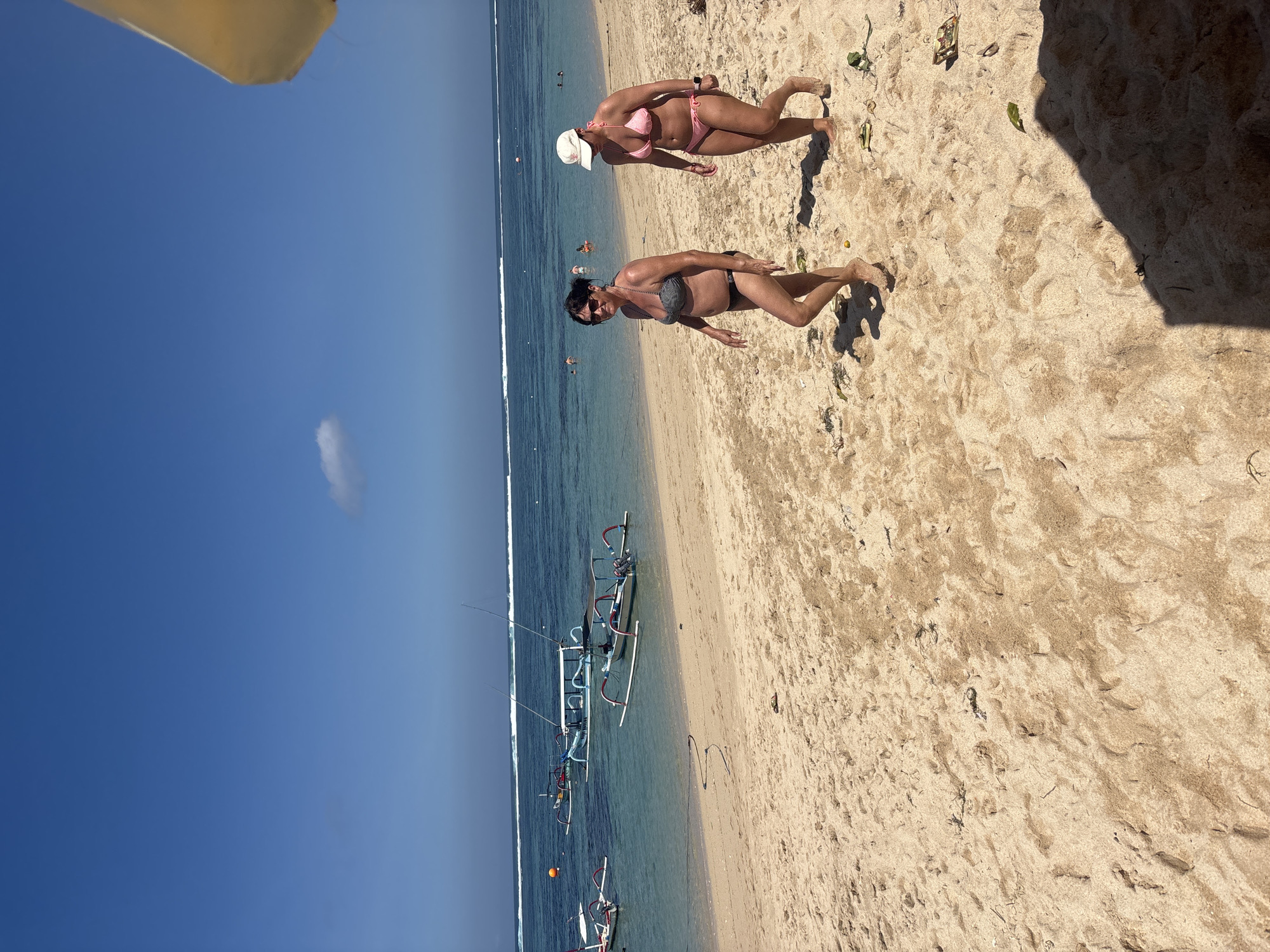 Two women walk along a sandy beach in Kuta Selatan, Indonesia. The woman on the left wears a dark grey and black bikini, sunglasses, and has dark hair. The woman on the right wears a pink bikini and a white baseball cap. The ocean is visible behind them, with several traditional outrigger boats anchored in the turquoise water. Gentle waves break on the horizon. The beach is covered in light-colored sand with some scattered debris. The sky is clear blue with a single, small white cloud. The sun casts shadows on the sand, indicating it is daytime.