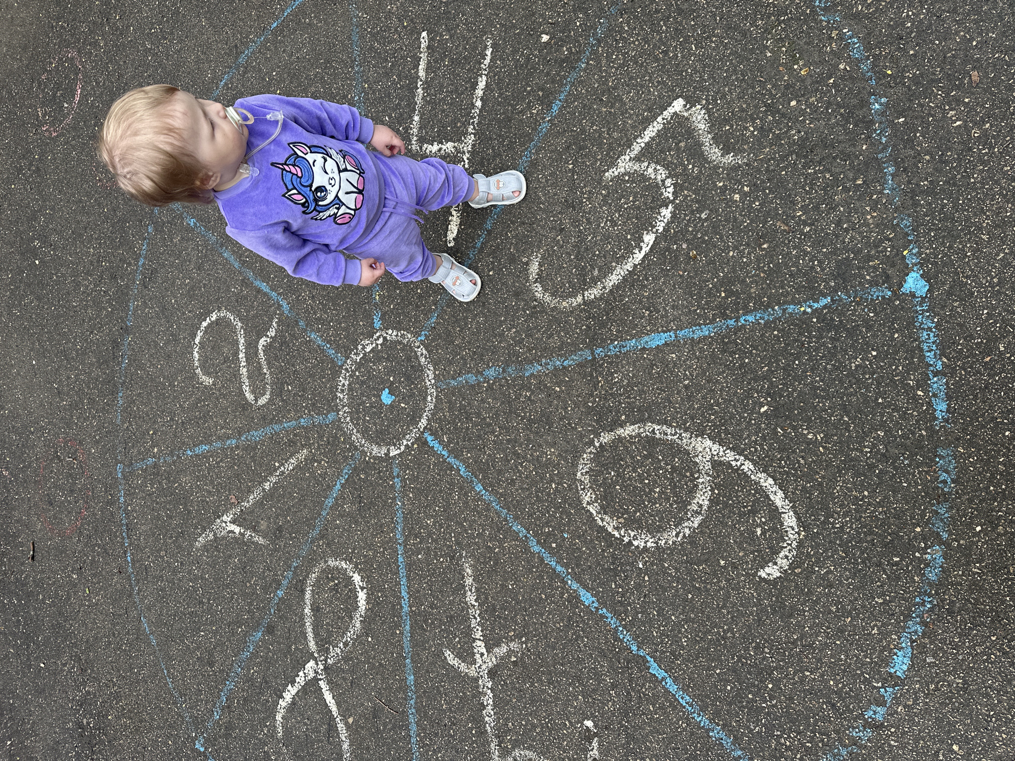 A small child wearing a purple sweatsuit with a unicorn on the front is standing on a gray asphalt surface with a clock face drawn on it in white chalk. The clock face has blue chalk lines radiating from the center. The numbers 4, 5, 6, 7, 8, 9, 10, and 11 are written around the perimeter of the circle. The location is likely outdoors on a playground or sidewalk. The city and country of the image is unable to be determined.