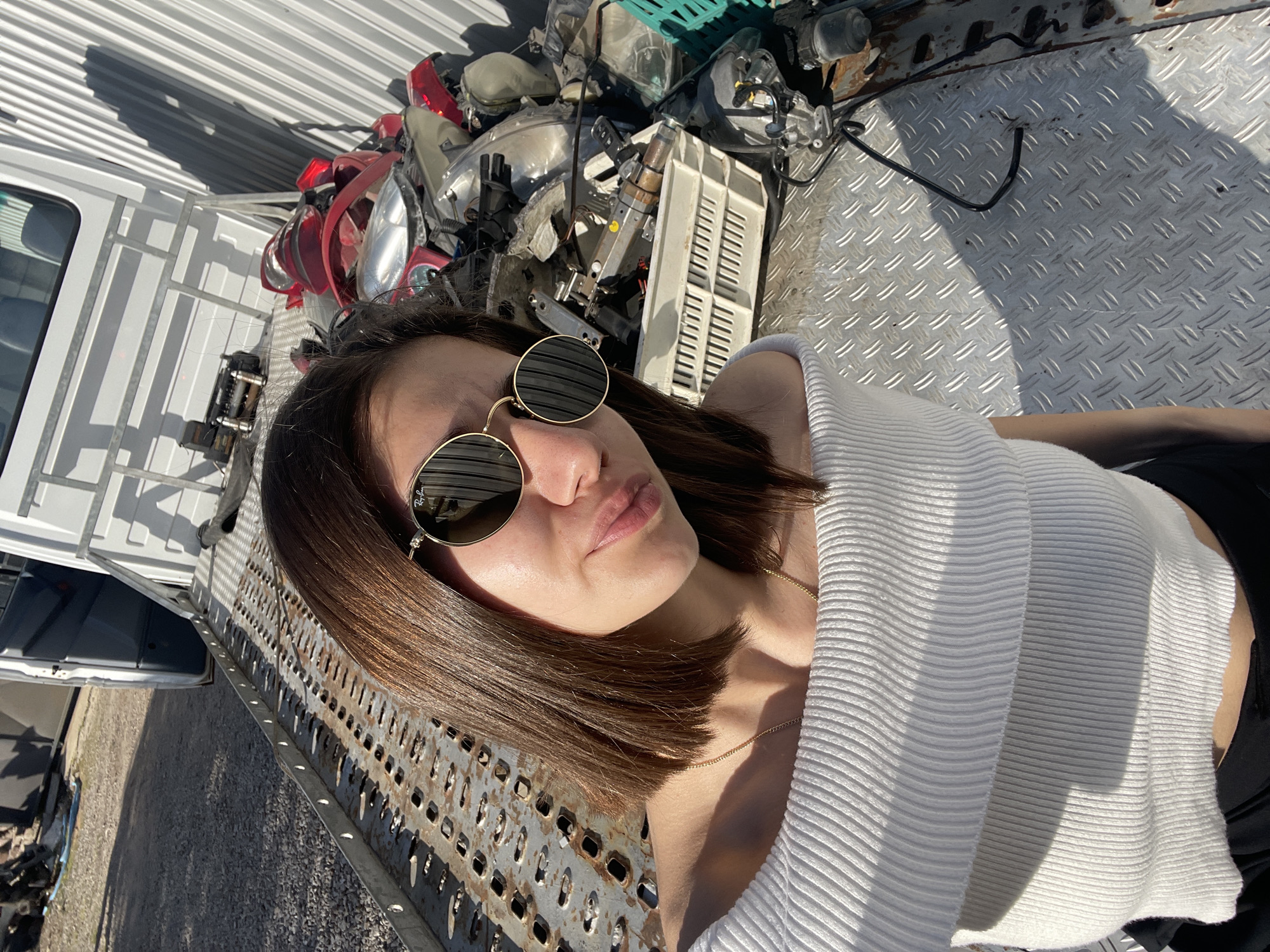 The image features a young woman lying on a textured metal surface, gazing towards the camera. She has short, straight, dark brown hair and wears round, gold-framed sunglasses with dark lenses, which reflect the brand name "Ray-Ban." Her lips are slightly pursed in a posed expression. She is dressed in a form-fitting, white, ribbed top and a thin gold chain necklace.
She rests on what appears to be the bed or ramp of a commercial vehicle, possibly a tow truck or flatbed. The surface directly beneath her includes a diamond plate pattern, and to her side, a perforated metal grate is visible. Behind her head, there is a large collection of miscellaneous automotive parts, including various headlights and red taillights, suggesting a setting like a junkyard, scrapyard, or auto repair facility. A white commercial vehicle with ridged side panels is partially visible in the background. The ground seen in the distance is gravel.
The scene is illuminated by bright, clear sunlight, indicating a daytime setting with strong shadows. The weather appears sunny with no visible clouds. The overall impression is a casual, posed portrait set within an industrial or automotive-focused environment.