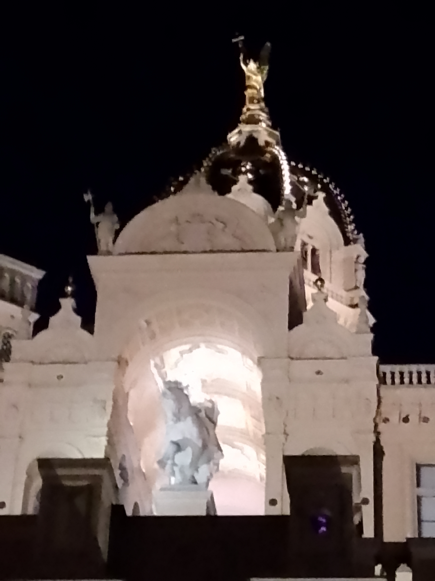 The image shows a night view of the Schwerin Cathedral in Schwerin, Germany. The cathedral features a prominent dome topped with a golden statue of an angel holding a cross. The white stone facade of the cathedral is illuminated, highlighting architectural details such as arches, statues, and decorative elements. In the foreground, a large white sculpture of a horse and rider is visible within an arched opening. The sky is dark and clear, suggesting it is nighttime. The overall impression is one of grandeur and religious significance.