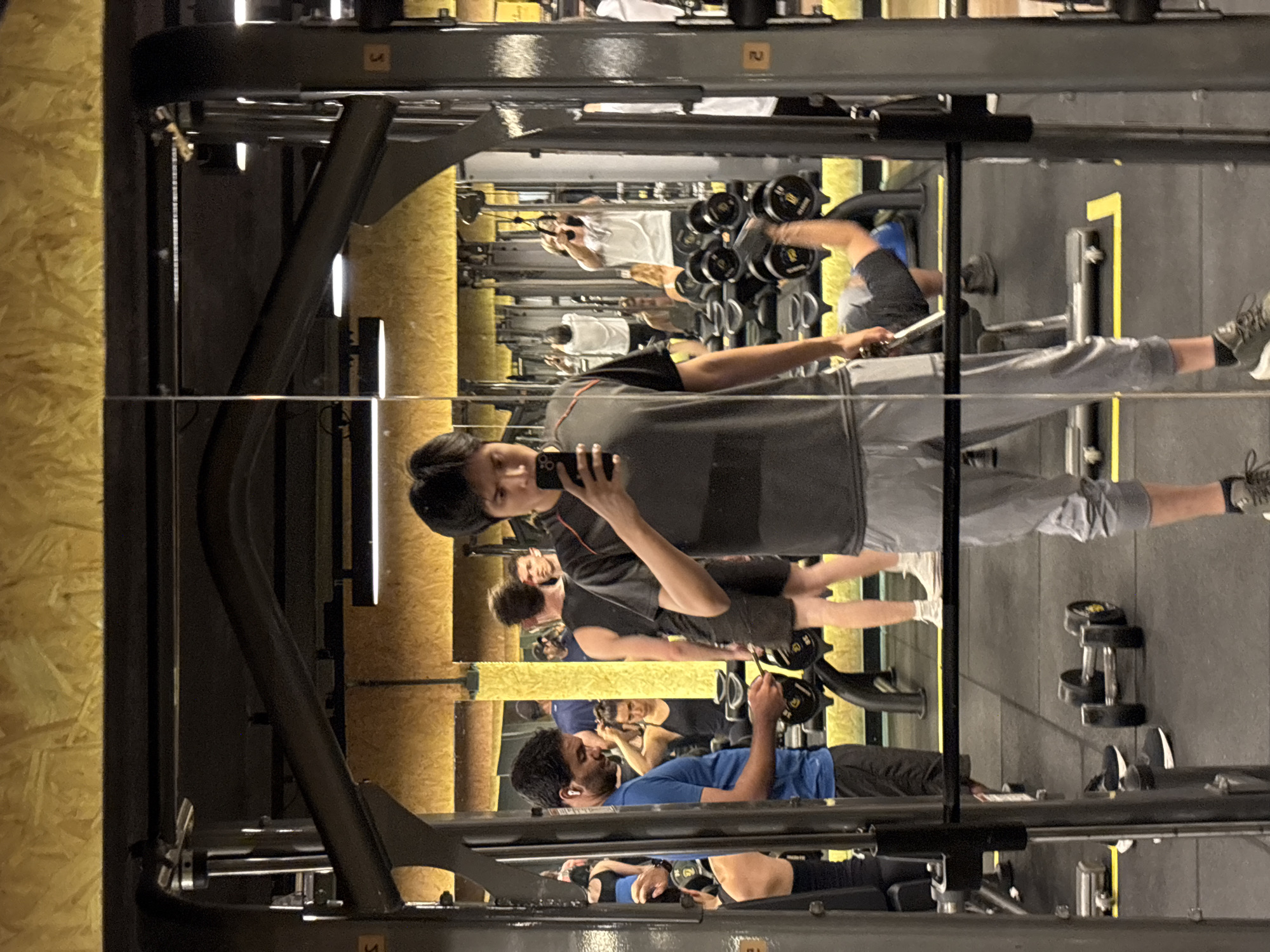 The image shows the interior of a gym, reflecting in a mirrored wall. Multiple people are present, engaged in various exercises. A man near the mirror is holding a smartphone, possibly taking a selfie. Another individual is working with dumbbells, while others are using gym equipment. The gym features weight racks, strength training machines, and free weights. Walls appear to be a combination of wood panelling and grey textured material. It is not possible to determine the specific location in São Paulo, Brazil from the image alone.