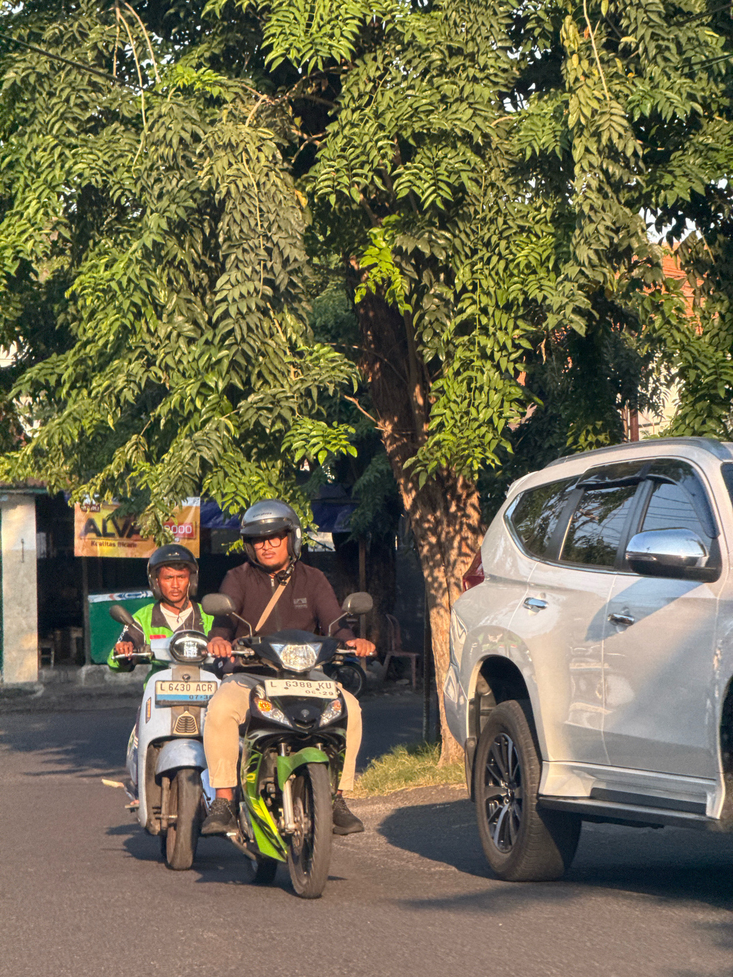 The image depicts a street scene in Surabaya, Indonesia, under a large, leafy tree. Two men are riding on a motorcycle. The rider in front wears a lime green jacket and a black helmet, while the passenger behind him wears a dark long-sleeved shirt, glasses, and a grey helmet. They are both facing forward, as if traveling down the road. Beside them, a white SUV is parked or driving slowly, its side and wheel prominently visible. In the background, there's a building with a banner that reads "ALVA" and some trees. The sunlight casts shadows from the tree onto the road and the SUV.