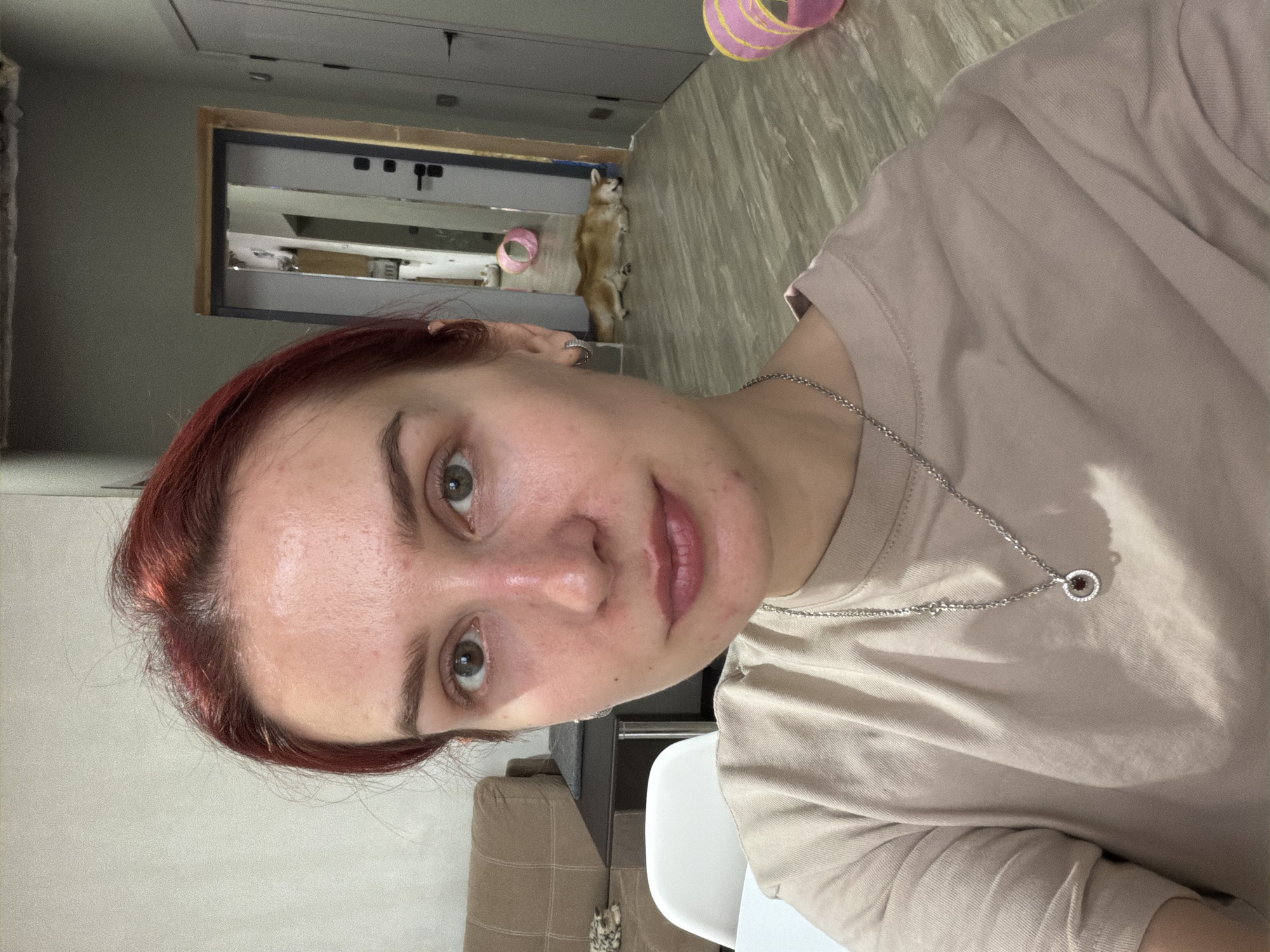 The image is an indoor selfie of a young woman, captured with a 90-degree counter-clockwise rotation. The woman, with reddish-brown hair and brown eyes, is looking directly at the camera with a slight smile. Her skin shows some visible blemishes. She wears a plain, light beige t-shirt and a silver chain necklace with a small, circular pendant featuring a red central stone surrounded by tiny white stones.
In the background, a small, light brown dog, possibly a Shiba Inu, is lying on its back on the floor, visible with its paws curled up. A pink and yellow pet toy is near the dog. The interior space features light-colored flooring, possibly wood or laminate, and light grey walls. A white chair and a section of a light brown sofa are also visible. A doorway or opening is present in the background, along with a rectangular mirror or reflective panel in a light wooden frame. The lighting suggests it is daytime. There is no visible text in the image. The location context for this scene is Chaikovsky, Russia.