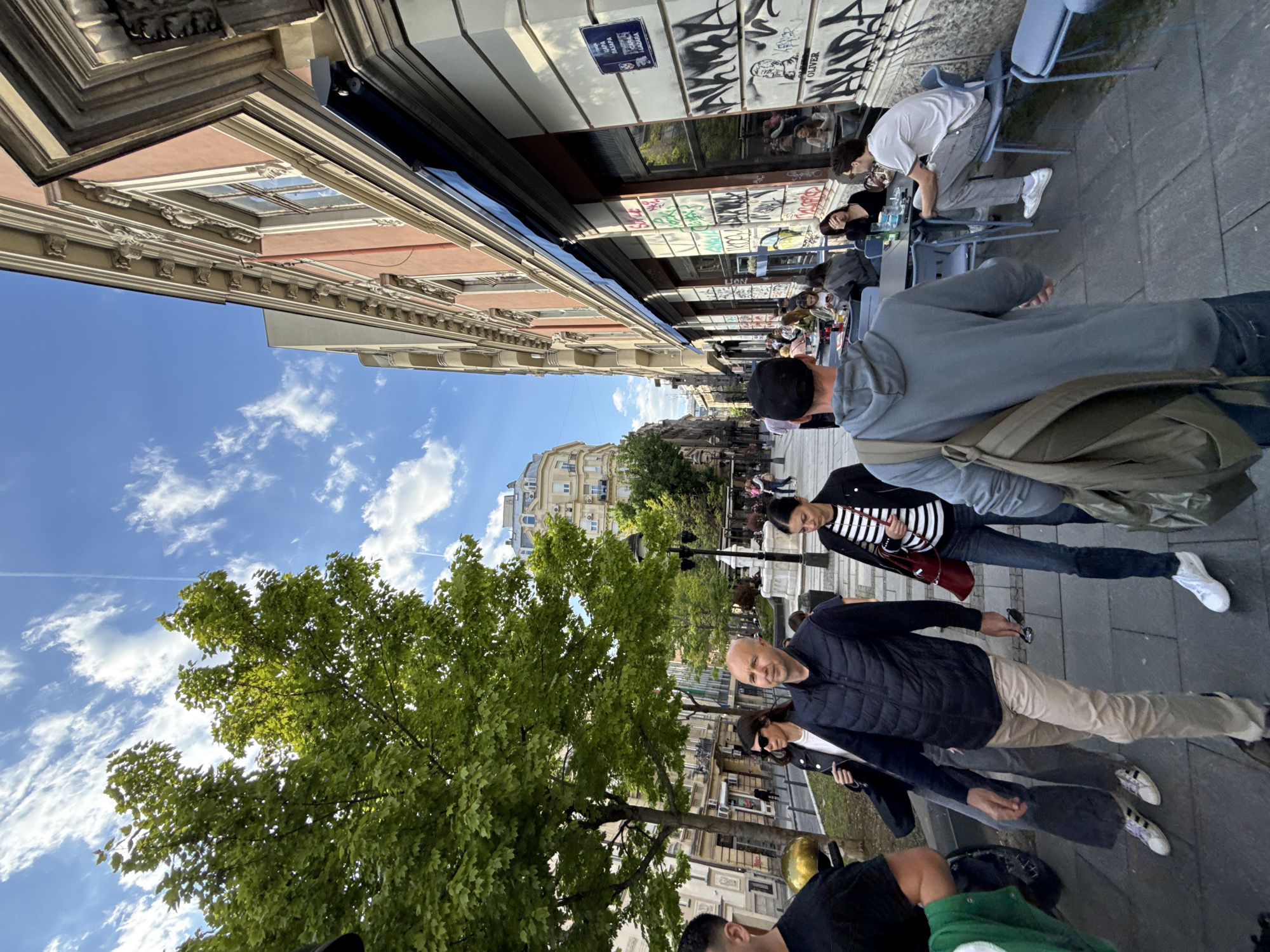 This image depicts a bustling street scene in Belgrade, Serbia, under a bright blue sky dotted with fluffy white clouds, indicating clear daytime weather. The perspective looks down a lively street flanked by buildings, trees, and pedestrians.
On the left side of the street, a multi-story building with ornate classical architecture features a reddish-pink upper facade and detailed cornices and window frames. A large, verdant tree with bright green leaves stands prominently in the foreground on this side, partially obscuring the building.
On the right side, across the street, a vibrant street-side cafe is active with patrons. Blue tables and chairs are visible on the sidewalk. The building housing the cafe has a dark awning, and the wall above it is heavily adorned with various graffiti tags, including the discernible name "OLIVER." A blue plaque on this building is visible, bearing text in Cyrillic, which appears to read "ГРАДСКА СКАЛА" (Gradska Skala).
Many people are present throughout the scene. In the immediate foreground on the right, the back of a person wearing a grey hoodie and carrying a tan messenger bag is visible. Further into the cafe area, a man in a white t-shirt and grey pants is seated backwards on a blue cafe chair. Another man in a dark vest and light-colored pants walks towards the viewer in the mid-ground on the left, while a woman in a striped shirt and jeans, carrying a red purse, walks away from the viewer. Other figures are scattered around the cafe and sidewalk, indicating a busy urban environment. A glimpse of a brass instrument, possibly a tuba, is visible at the bottom center. The atmosphere is that of casual street life in a vibrant city.