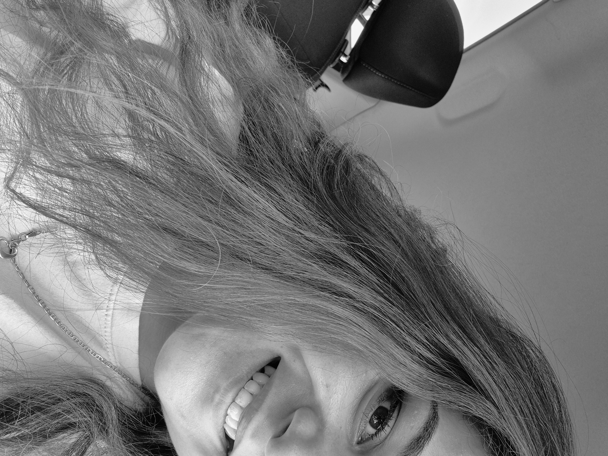 This black and white, low-angle photograph captures a close-up portrait of a person, likely female, inside a car in Sofia, Bulgaria. The subject's head is tilted, with long, straight hair flowing and spreading across a significant portion of the frame, suggesting a relaxed or reclined pose.
Only parts of the face are visible: one eye, part of the nose, and a wide, open-mouthed smile revealing bright teeth. The subject's expression appears cheerful and engaged, with the visible eye looking directly towards the viewer. She is wearing a light-colored top, and a delicate chain necklace with a small heart-shaped pendant is visible near her left shoulder.
The setting is an interior of a car, indicated by the dark, upholstered headrest visible in the upper right corner and the light-colored ceiling or roof of the vehicle occupying the background. The unique, almost inverted perspective creates an intimate and dynamic composition. There is no visible text in the image.