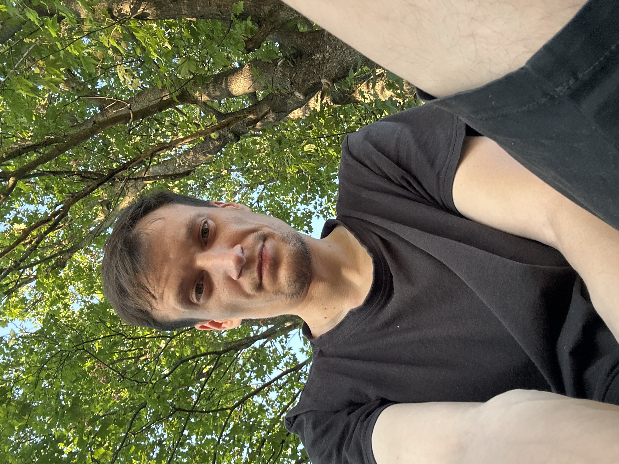 A selfie is taken from a low angle, looking up at a man. He is wearing a black t-shirt and black shorts. He has short, dark brown hair and light brown eyes. His skin is fair. The background is a tree with green leaves. The image was taken in Makiv, Ukraine, on a sunny day.