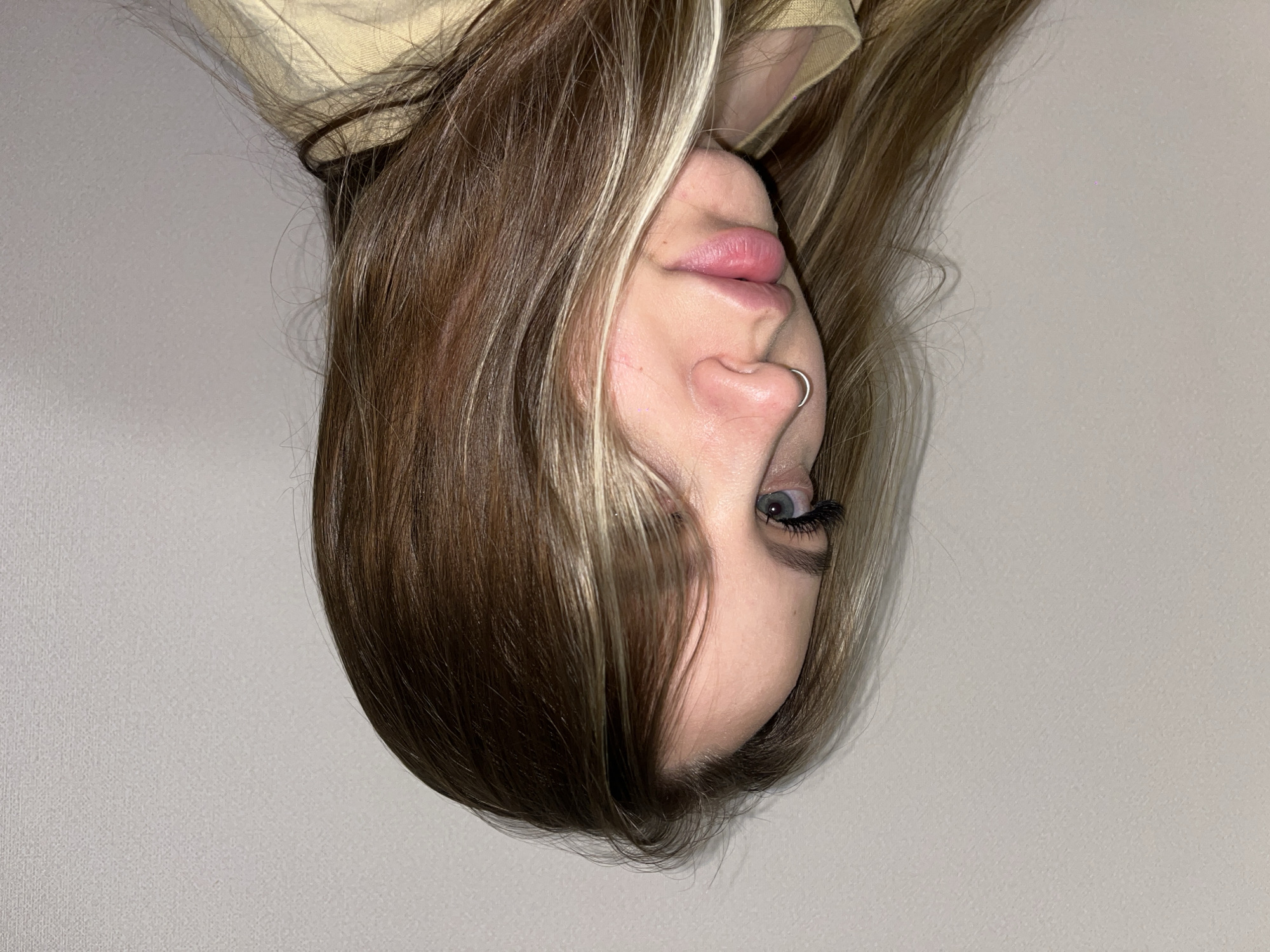 A close-up shot of a person's face with light brown hair and blonde highlights, with the image rotated upside down. The person has a nose ring, visible eyelashes, and light-colored eyes. The background is a plain light gray. The location is not discernible, and there is no information to suggest the image was taken in Krasnodar, Russia.