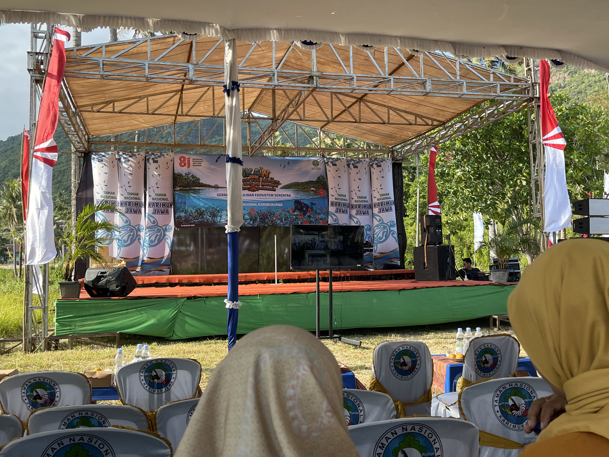 The image shows an outdoor stage set up for an event. The stage has a tan canopy supported by a metal frame. Red and white flags are displayed on either side of the stage. Several vertical banners with blue and white designs hang behind the stage. A large screen is positioned in the center of the stage. In front of the stage, rows of white chairs with blue and white logos are set up, some covered with plastic. In the foreground, the backs of two individuals wearing headscarves are visible, one in a yellow headscarf and the other in a patterned beige headscarf. Water bottles and fruit are placed on a blue table between some of the chairs. The setting appears to be a grassy area with trees and hills in the background. The event is likely taking place in Kemloko, Indonesia, as suggested by the banner text which includes "KARIMUN JAWA," a well-known destination in Indonesia.