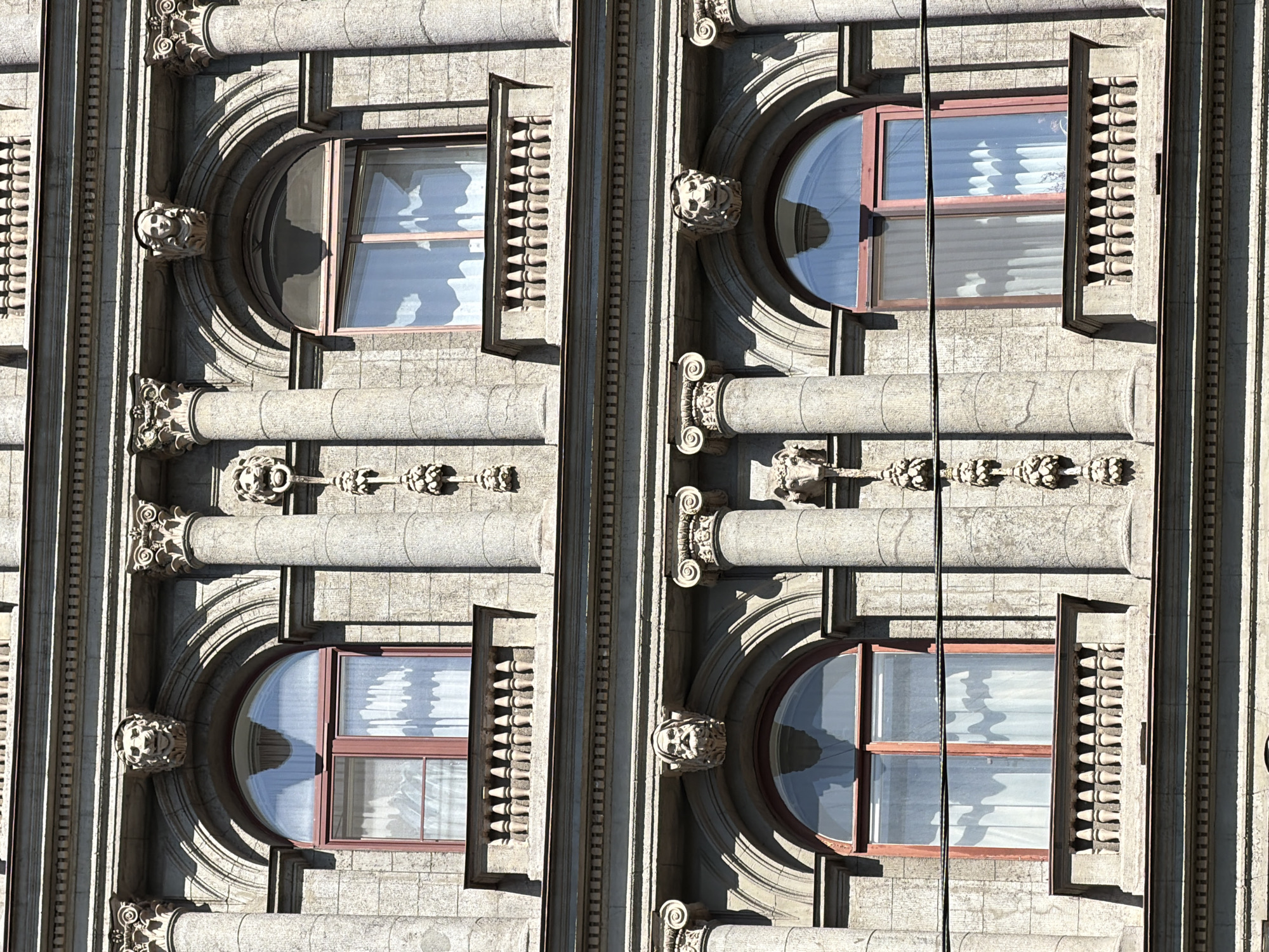 The image captures a detailed section of a building facade, showcasing ornate classical architectural elements. The stone-clad structure features two vertical segments, each comprising an arched window on the left and a rectangular, vertically oriented decorative panel on the right.
The arched windows have multi-pane sashes with a reddish-brown frame, reflecting a bright sky and interior white curtains. Below each arched window, a prominent carved stone grotesque or lion's head adorns the facade, serving as a decorative bracket. These are flanked by short, stout columns with ornate, possibly Ionic, capitals.
To the right of each arched window is a tall, narrow rectangular opening filled with a series of conical or pointed decorative elements, resembling a stylized balustrade or a dentil pattern. The entire facade is constructed from grey, textured stone blocks, exhibiting a substantial and historic appearance.
Strong sunlight illuminates the building from the left, casting deep shadows that highlight the three-dimensional relief of the carvings, columns, and recessed window frames. This suggests it is daytime, likely late morning or early afternoon, under clear skies. A thin, dark cable or wire runs vertically down the right side of the image, obscuring a small part of the facade. The architectural style, with its rich detailing and classical influences, is consistent with buildings found in Saint Petersburg, Russia. There is no visible text.