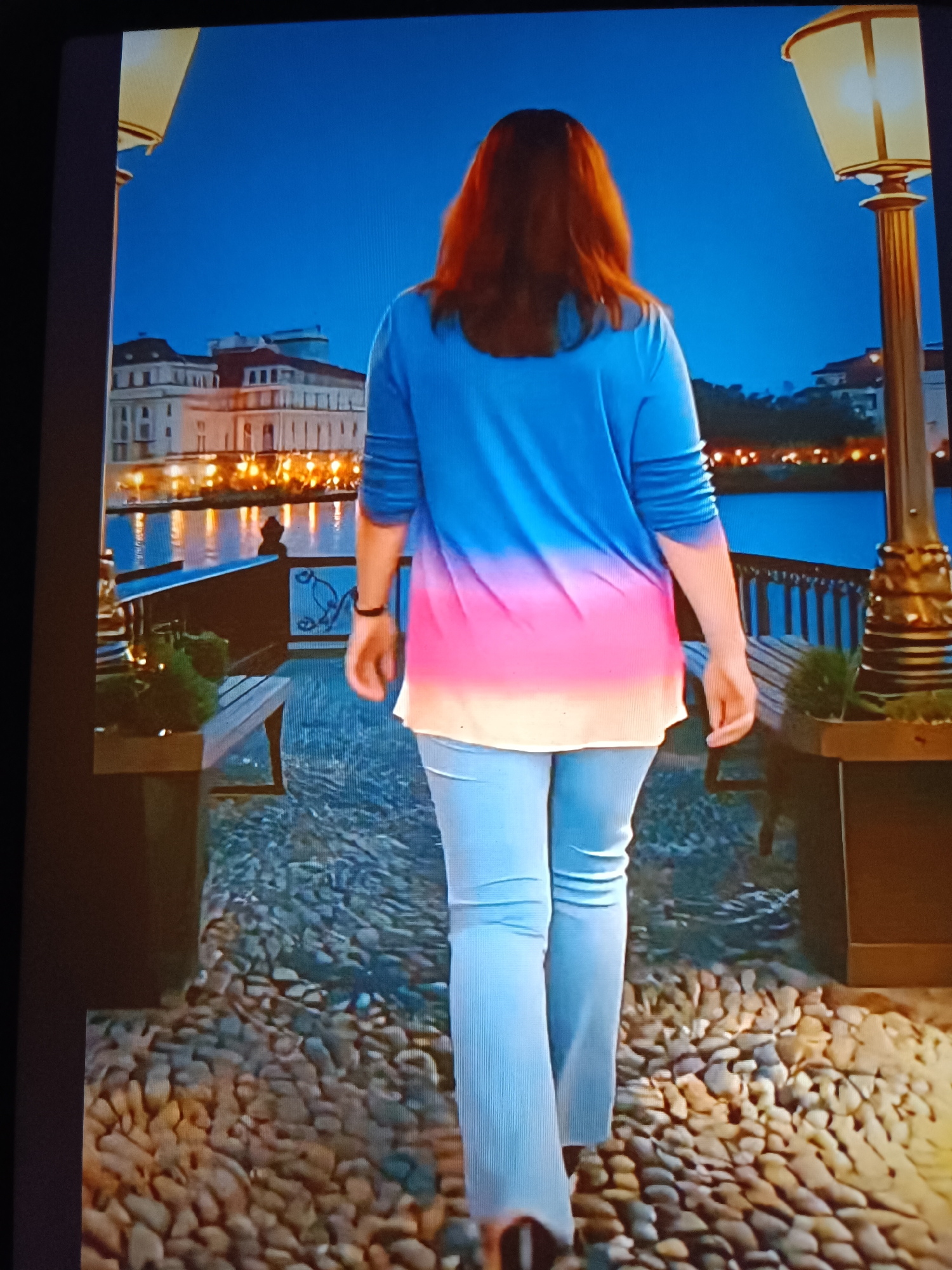 The image captures a person walking away from the viewer on a cobblestone path in the city of Tebras, Latvia, during the evening or night. The person, identified as female, has medium-length reddish-brown hair and is observed from the back. She is wearing a long-sleeved top with an ombre pattern, transitioning from blue at the shoulders to pink in the middle, and off-white at the hem. Her lower attire consists of light blue trousers or jeans. The path is composed of various-sized, rounded cobblestones or pebbles. The scene is bordered by two ornate, illuminated lampposts; one partially visible on the left and a more complete one on the right. In the midground, a dark railing with decorative elements separates the path from a body of water, which reflects numerous golden lights originating from buildings on the opposite bank. The background features a dark blue sky above several brightly lit, multi-story buildings exhibiting traditional architectural styles across the water.