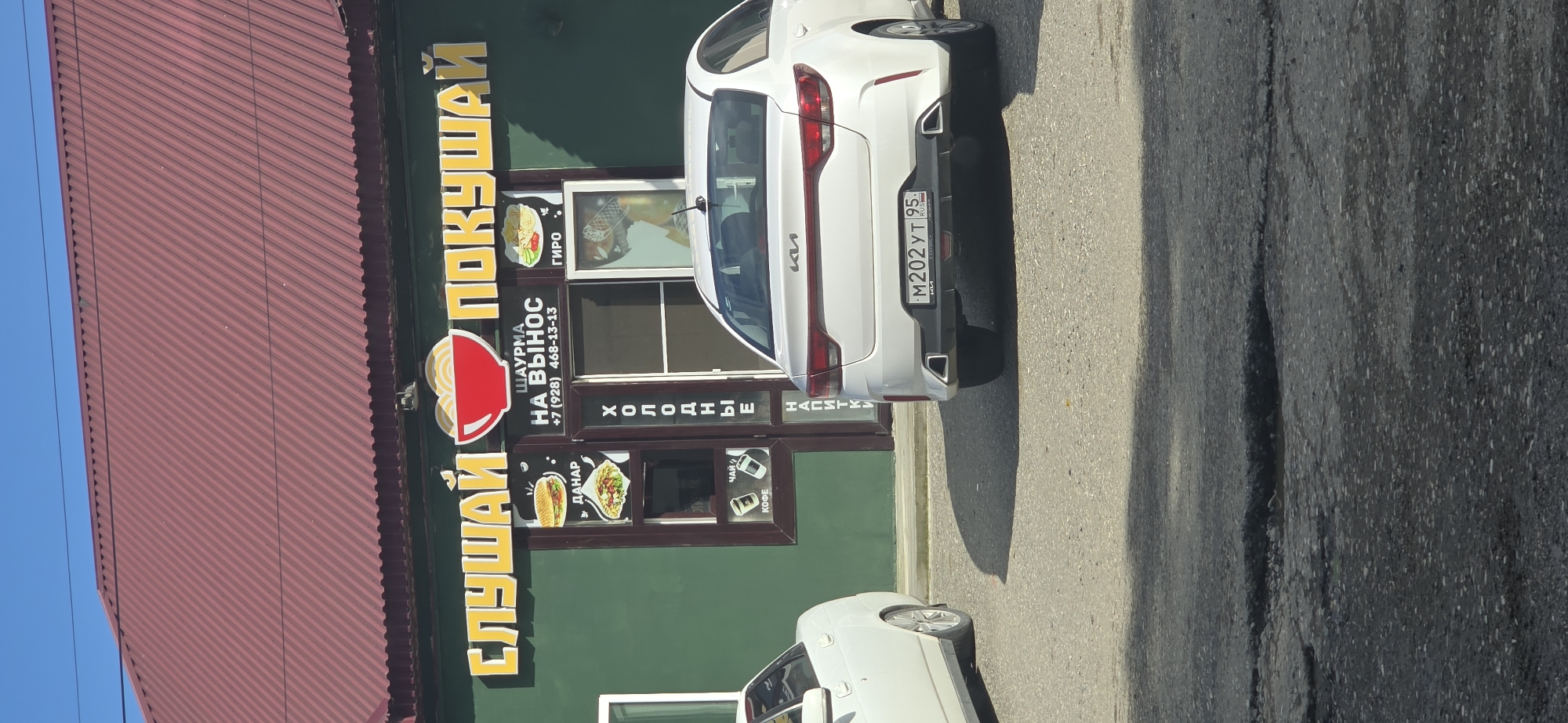 The image depicts a street scene with a food establishment named "Слушай Покушай" ("Listen and Eat") in Grozny, Russia, based on the license plate (95) of the white Kia car parked in front. The sign advertises "Шаурма на вынос" ("Shawarma to go") along with other food items like gyro, danar, and drinks. The phone number "+7 (928) 468-13-13" is also displayed. The building is green with a red roof. The car's license plate reads "М202УТ95". The street is paved.