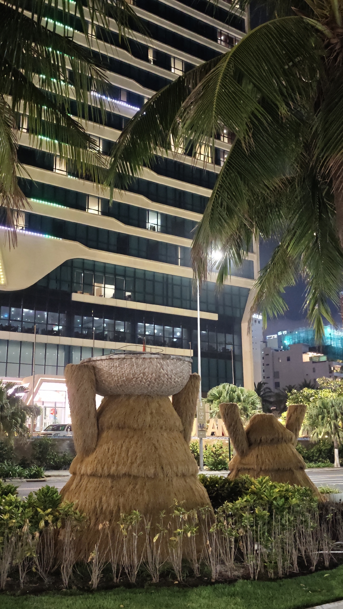 The image captures a night scene in Đà Nẵng, Vietnam. In the foreground are two large, conical sculptures constructed of straw or similar fibrous material. The larger sculpture on the left supports a round, woven basket on top. The sculptures are surrounded by low shrubs and greenery. Behind the sculptures, a modern, multi-story building with glass windows and horizontal white bands is visible. Palm trees frame the scene on either side, with their fronds partially obscuring the building. A street with faint car lights and a street light can be seen in the background. The overall lighting suggests an urban environment with streetlights and building illumination.