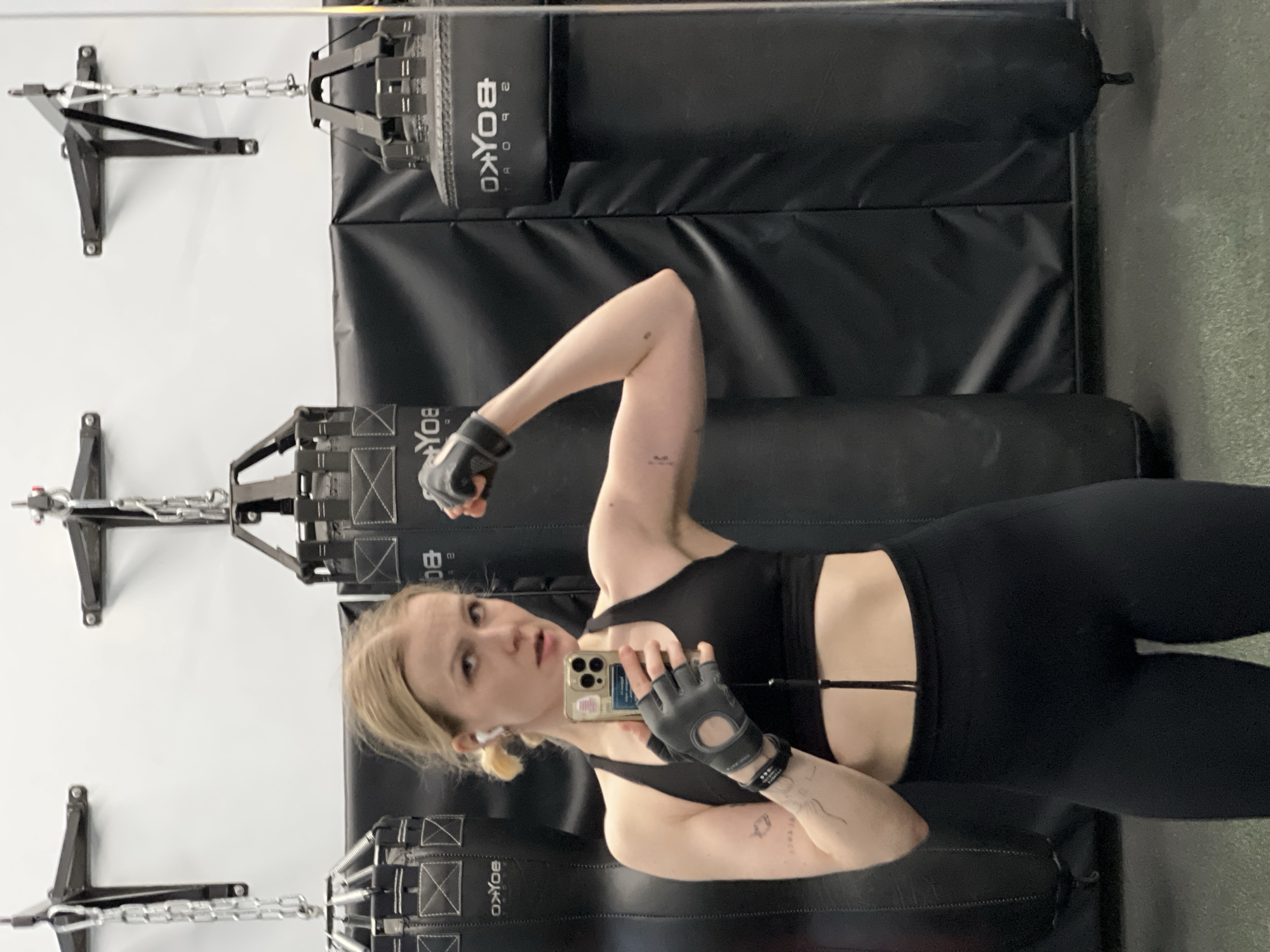 A fair-skinned woman is taking a selfie in what appears to be a gym. She is wearing black workout clothes, fingerless gloves, and has blonde hair pulled back in a ponytail. Her right arm is flexed. She is in front of a black padded wall with two heavy punching bags suspended by chains. The wall and punching bags have "BOYKO" printed on them. The location cannot be determined to be Khimki, Russia, as the image provides no evidence to suggest that.