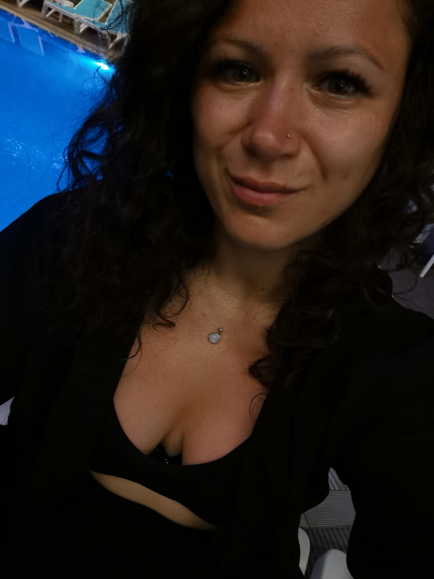 A woman with dark, curly hair and a nose stud poses for a selfie near a brightly lit blue swimming pool. She is wearing a black top that reveals cleavage and a delicate silver necklace with a round pendant. The background shows the edge of the pool with lounge chairs and a hint of a tiled walkway. The lighting suggests it is evening or nighttime. The location is identified as Alanya, Turkey, and the setting appears to be an outdoor pool area at a resort or hotel.