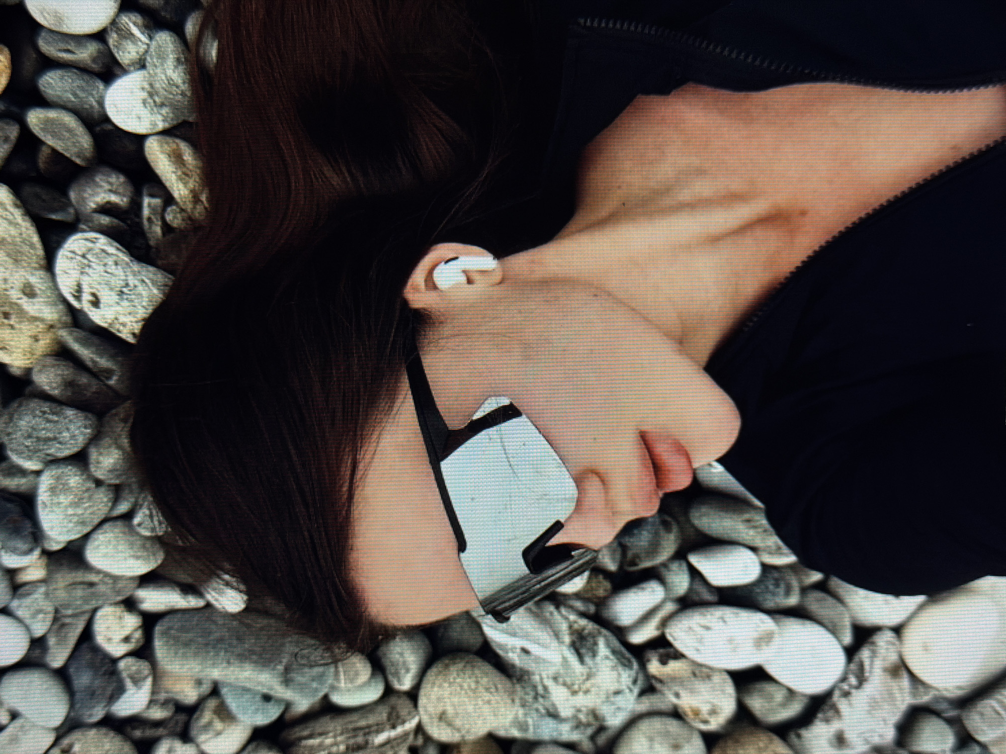 A woman with dark hair, wearing large, reflective sunglasses and a white wireless earbud, lies on her back amidst a bed of grey and white pebbles. She is wearing a black zip-up jacket. The scene appears to be outdoors, possibly near water. The image is a close-up shot, focusing on the woman's face and upper body. The location in the city of Sirius, Russia is not specified within the image or its context.