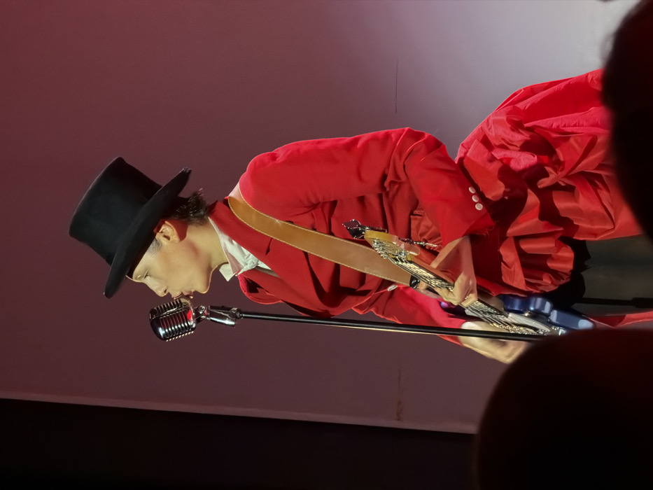 On a stage in Moscow, Russia, a performer is depicted mid-action. The individual wears a black top hat, a red velvet-like tailored jacket over a white ruffled collar, and a voluminous red pleated lower garment, with black trousers visible beneath. They hold a blue electric bass guitar, secured by a brown leather strap, with hands positioned for playing. The performer's lips are pursed towards a chrome vintage-style microphone on a stand. The background is a stark, reddish-pink illuminated stage wall with a lighter vertical element on the left. The blurred tops of audience members' heads are visible in the immediate foreground, indicating a live performance setting.