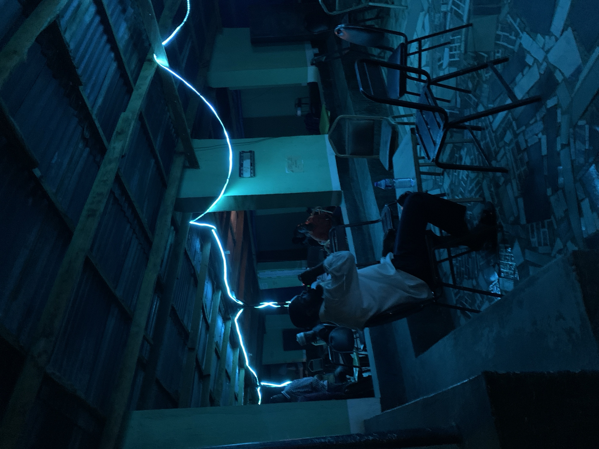 The image captures a dimly lit indoor space, dominated by shades of blue and teal from artificial lighting. A vibrant, glowing blue-cyan light strip runs diagonally upwards along the left side of the frame, illuminating the surrounding structures.
In the mid-ground, a person wearing a white shirt and dark trousers is seated on a chair, facing away from the camera. A plastic water bottle rests on a surface nearby. In the background to the right, several chairs are stacked or arranged on what appear to be tables. Further down the illuminated corridor on the left, another person in a striped shirt is partially visible, seated on a low surface.
The setting features a corrugated metal wall on the left, reinforced by visible wooden beams. Opposite this, the walls are painted in a teal hue, with concrete structures and sections of patterned flooring. An unreadable framed notice or sign is visible on a teal wall. The environment appears to be a functional, possibly communal area or corridor, likely at night or in a deeply shaded interior due to the dominant artificial light source. The scene is located within or near the New Stadium, Nigeria. No specific event beyond people sitting is clearly depicted, and no text is legibly visible.