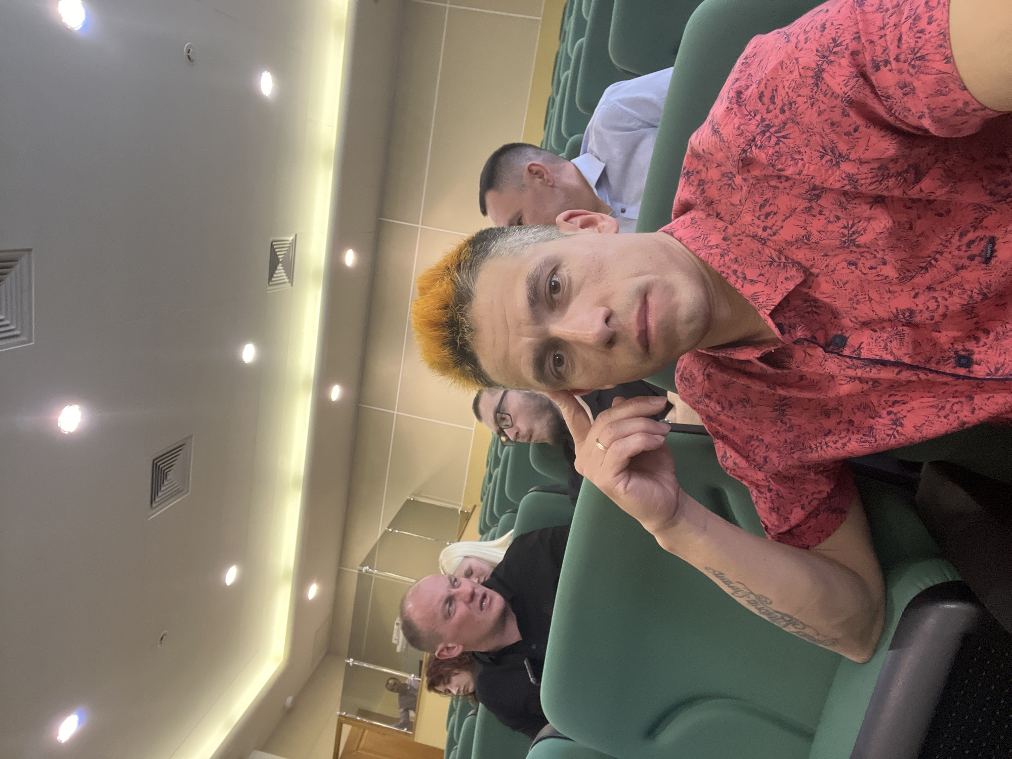 Location: Likely an auditorium or conference room in Izhevsk, Russia.
Scene: A selfie taken by a man with dyed orange hair, wearing a red patterned shirt. Several people sit in rows of green upholstered chairs behind him. One man wears a light blue shirt. A bald man wearing a black shirt is visible further back. Overhead are ceiling lights and ventilation grilles. The picture is taken at an angle.