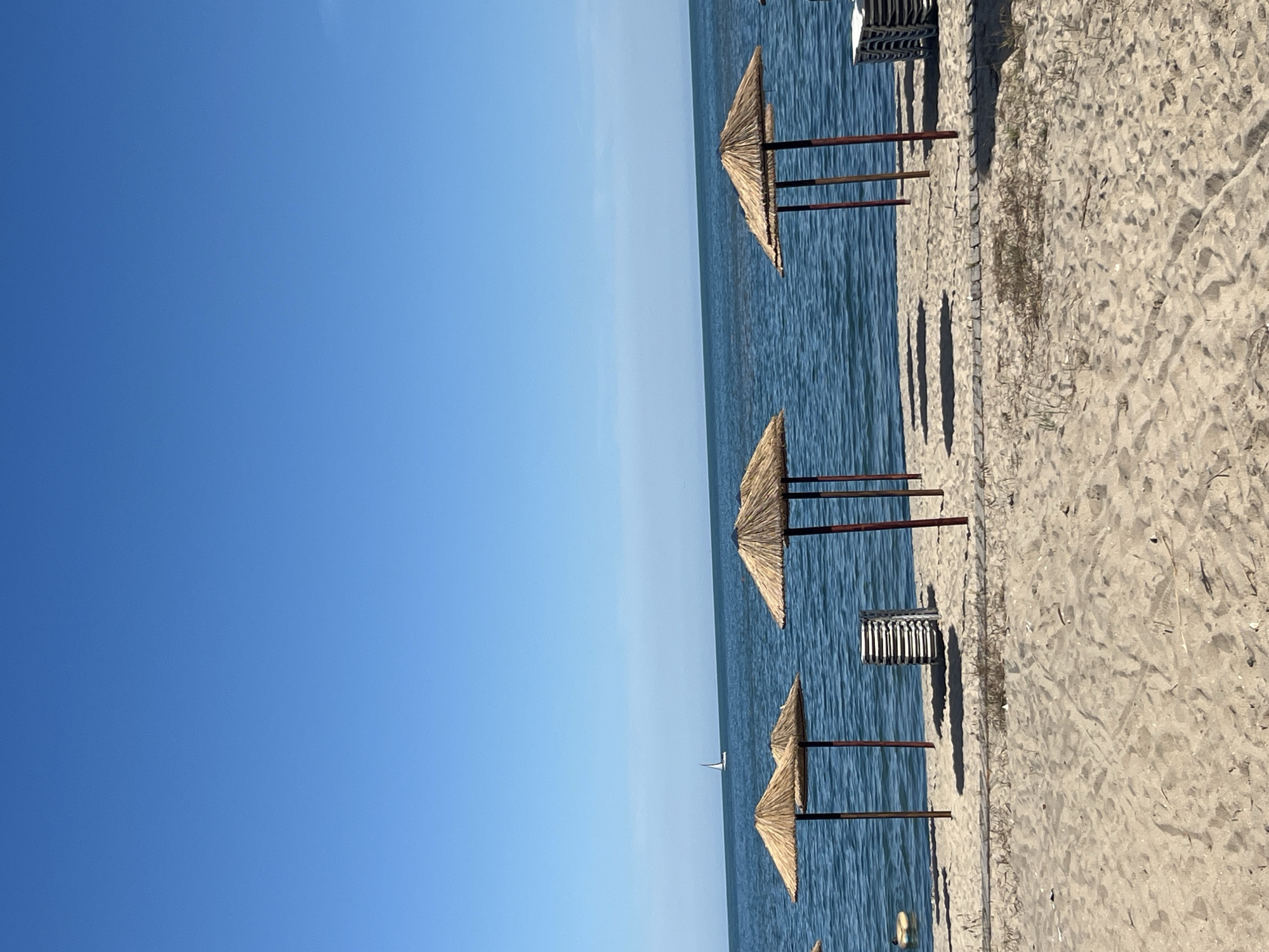 The image depicts a sandy beach with several straw umbrellas casting shadows on the sand. The sea is calm and blue, with a single sailboat visible in the distance. The sky is clear and bright blue. In the foreground, the sand is textured with footprints and scattered dry grass. To the left of the frame, a stack of white lounge chairs is visible near a straw umbrella. Further to the left, a yellow inflatable object floats in the shallow water. The scene suggests a tranquil beach setting, likely during a sunny day. The general setting is a beach in Varna, Bulgaria.