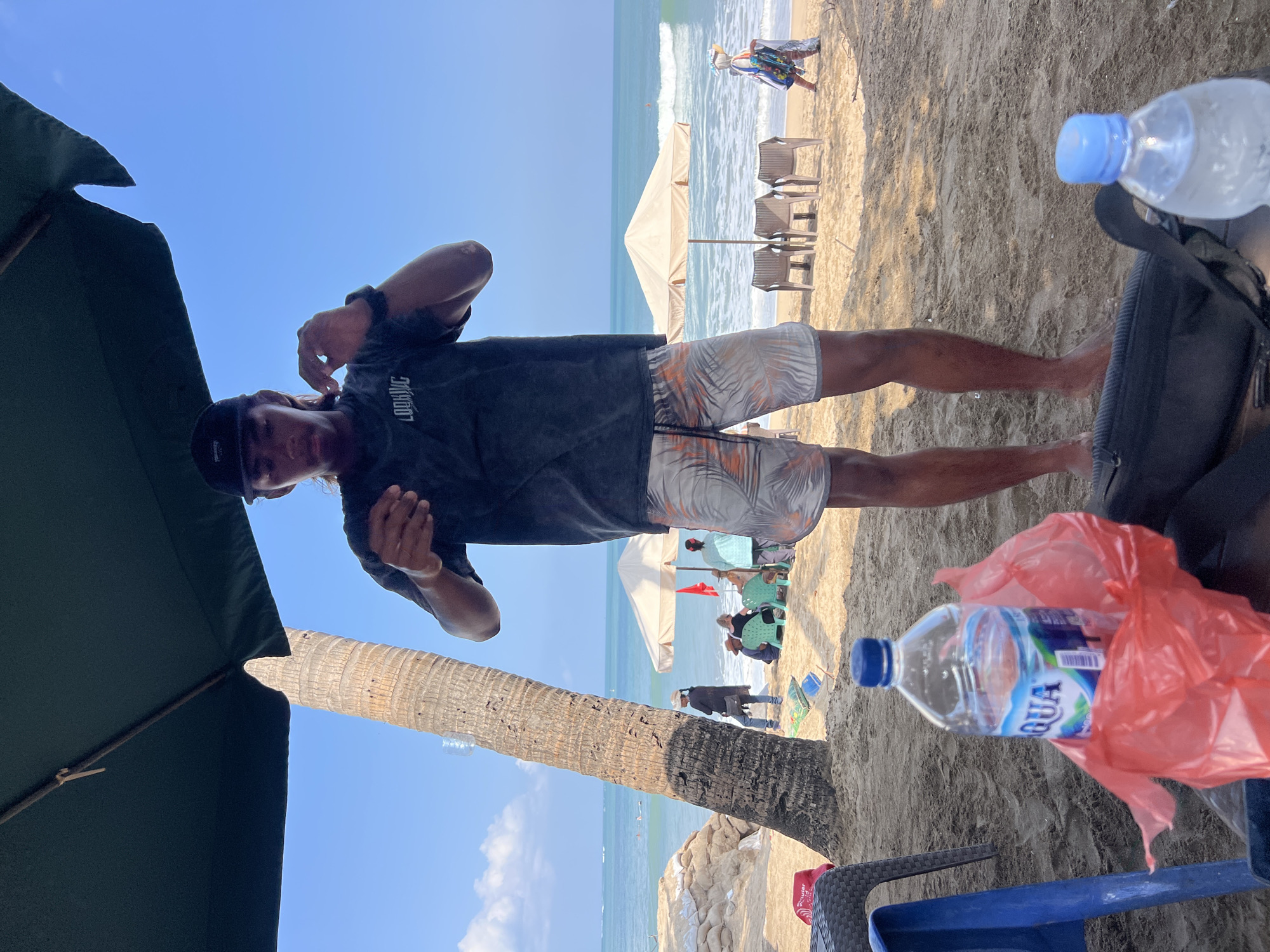 A young man wearing a black baseball cap and a dark t-shirt with the word "LOOKING" printed on it, and patterned shorts stands on a sandy beach in Kuta, Indonesia. He is facing the camera and appears to be adjusting something near his ear with his right hand. In the foreground, a blue plastic chair and a bottle of Aqua water are visible on the sand. Behind him, a large palm tree trunk stands to the left, and the ocean stretches out to the horizon under a clear blue sky. Several beach umbrellas and chairs are set up further down the beach, with a few people visible in the distance. The environment suggests a sunny day at the beach.