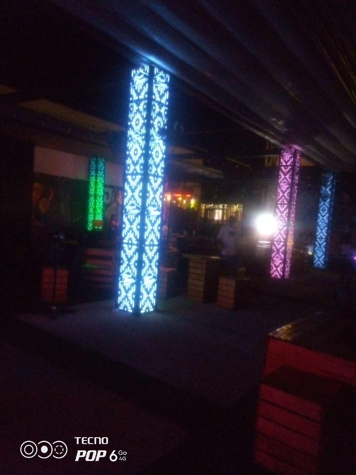 The image depicts a nighttime scene in an outdoor or semi-outdoor venue, possibly a bar or restaurant, in Howayi, Nigeria. Prominent features are tall, illuminated pillars with intricate, repeating patterns of light, primarily in blue and pink hues. In the background, there are glimpses of other structures, including what appears to be a building with windows emitting warm light, and some wooden crates or seating areas. A person is partially visible in the mid-ground, silhouetted against a bright light source. The overall ambiance is dark, with the illuminated pillars being the main source of light and color, suggesting a festive or entertainment-oriented setting. The ground appears to be paved or a dark, solid surface. The bottom right corner of the image contains a watermark with "TECNO POP 6 Go 4G".