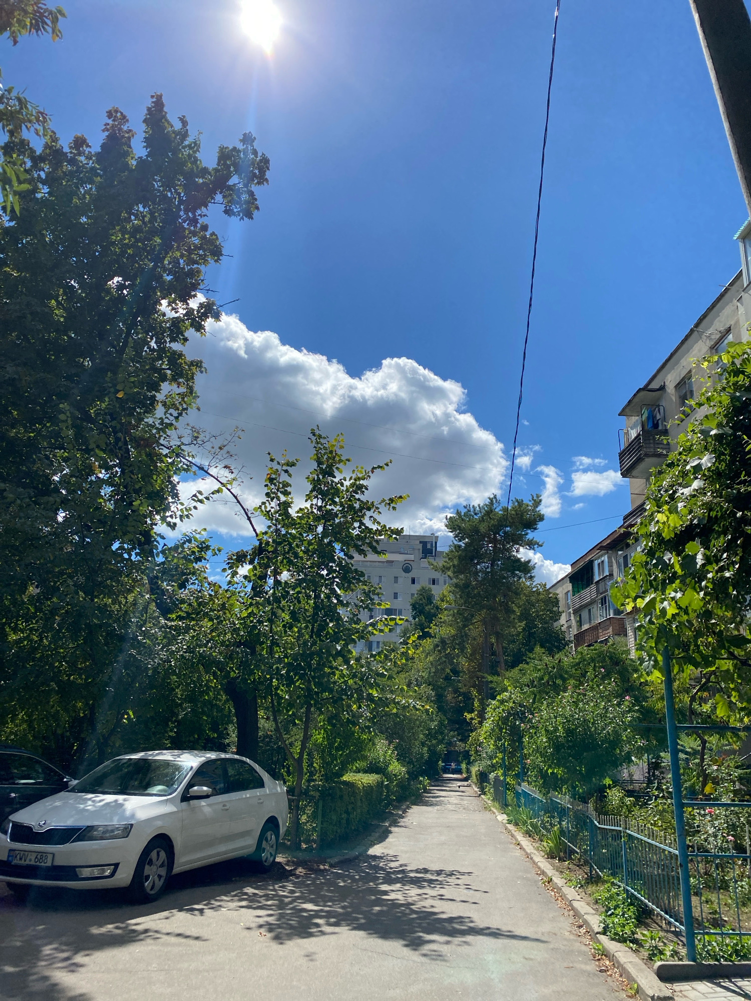 A white Skoda Rapid sedan is parked on the left side of a paved walkway in a residential area of Chișinău, Moldova. The car, with license plate KWV 688, is angled towards the viewer. To the right of the walkway is a blue metal fence, beyond which is a garden with various plants and flowers. In the background, multi-story buildings, characteristic of Soviet-era architecture, are visible, interspersed with trees. The sky is blue with scattered white clouds, and the sun is shining brightly, casting shadows on the walkway. A thin power line stretches across the upper portion of the image.