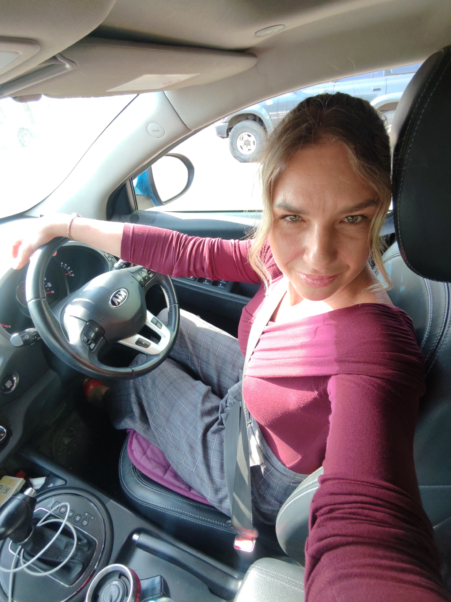 The image depicts a woman sitting in the driver's seat of a Kia car. She is wearing a maroon, off-the-shoulder top and gray pants with a plaid pattern. The steering wheel is black with the Kia logo in the center. She is wearing a gold bracelet. The background shows another vehicle, possibly a Toyota Land Cruiser. The location is unspecified, and there is no information to suggest that the location is Petropavlovsk-Kamchatsky, Russia.