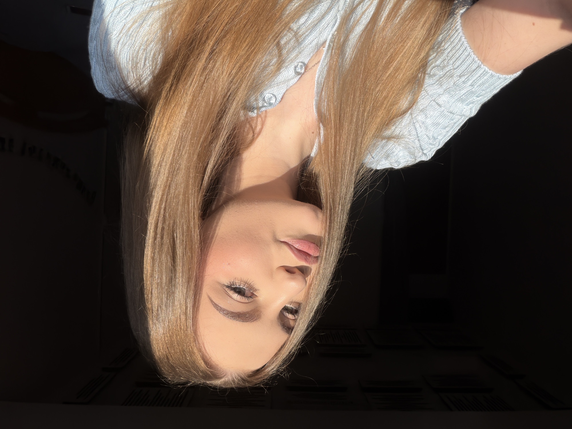 A young woman with long, straight, honey-blonde hair is captured in a close-up, front-facing shot. She is wearing a light blue, collared, button-up knit top. The woman's face is turned slightly to her left, with her head tilted. She is looking directly at the camera with full lips, painted with a light pink lipstick, and her eyes are open. Her makeup includes defined eyebrows, eyeliner, and mascara, with a touch of blush on her cheeks. The lighting suggests she is near a window, with sunlight illuminating the right side of her face and hair, casting shadows on the left. The background is dark, with some indistinct shapes suggesting an indoor setting. The image does not provide any specific clues to its location in Dnipro, Ukraine.