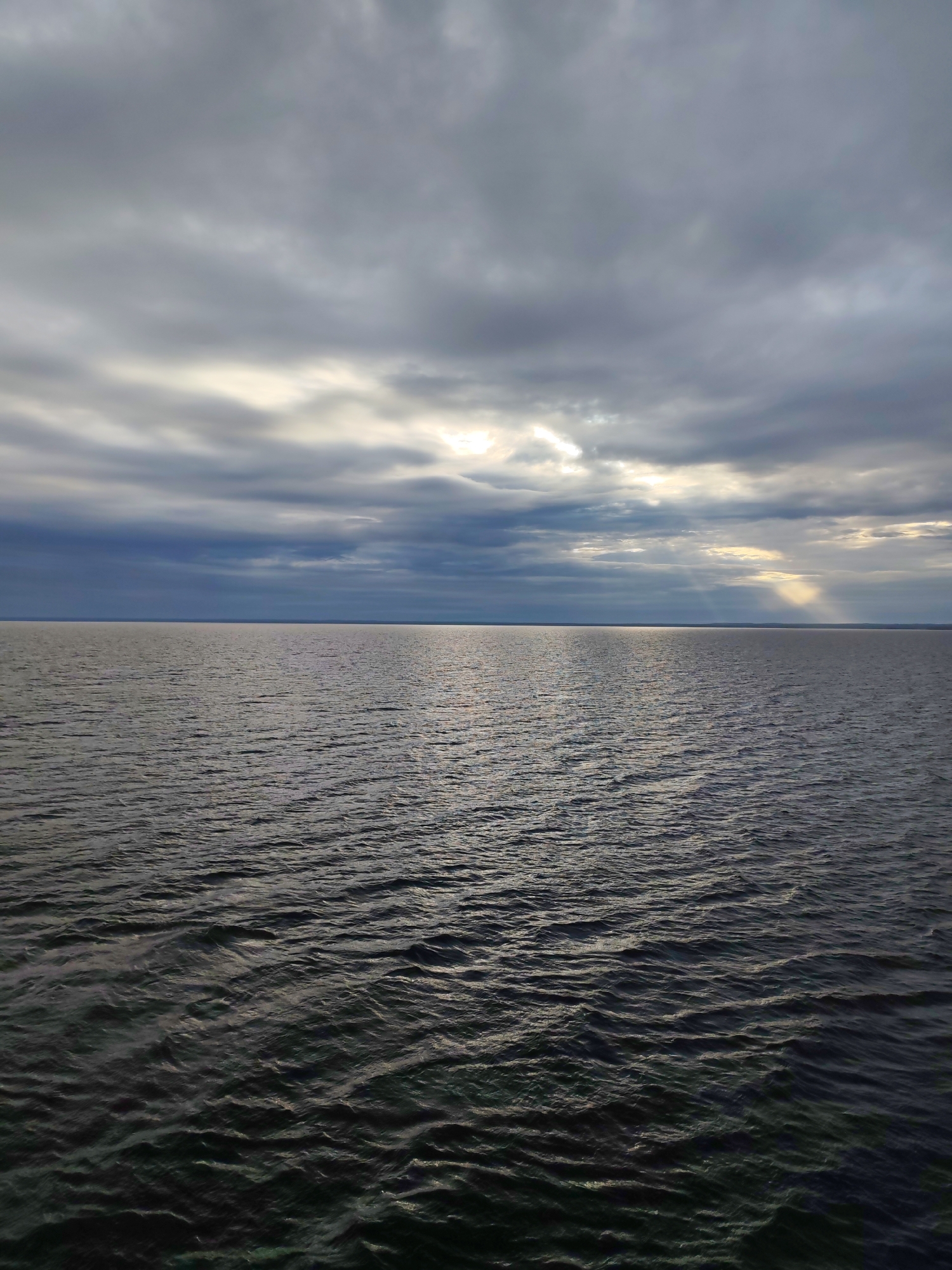 This media file captures an expansive natural landscape featuring a vast body of water under a dramatic sky. The scene is dominated by the dark, textured surface of the water, which occupies the lower two-thirds of the frame, showing numerous small ripples and waves reflecting subtle light. The water extends to a distant, straight horizon line that stretches across the entire width of the image.
Above the horizon, the sky is heavily overcast with a complex arrangement of grey and dark blue clouds. Breaks in the cloud cover allow beams of sunlight, known as crepuscular rays, to pierce through and illuminate sections of the sky and potentially the distant water surface, particularly on the right side of the frame. The overall lighting suggests either late afternoon or early morning, with the sun partially obscured but its presence still making an impact.
There are no visible people, specific objects, or discernible activity taking place, emphasizing the raw, unpopulated nature of the environment. The image conveys a sense of calm vastness, with the turbulent sky adding a dynamic element to an otherwise serene watery expanse. Given the location context of Kuzaranda, Russia, this is likely a view of the White Sea or a large lake in the region. No text is visible in the image.