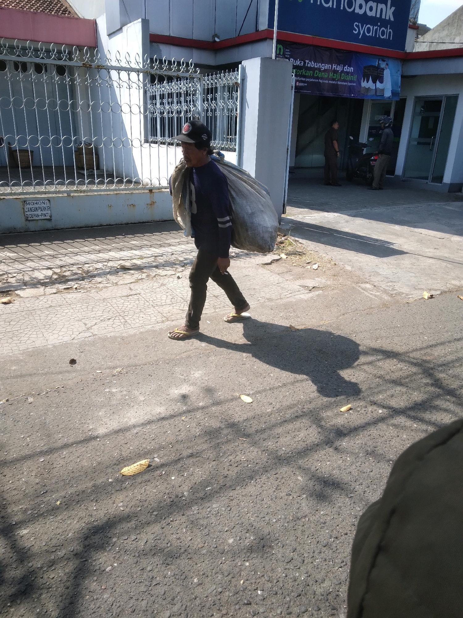 A man walks along a street in Purwokerto Kulon, Indonesia, carrying a large sack of what appears to be recyclables or trash over his left shoulder. He is wearing a dark blue long-sleeved shirt, dark pants, and yellow flip-flops. A black baseball cap is on his head. The man is casting a long shadow on the asphalt road, which is textured with small stones and has some scattered leaves and debris. Behind him, a white metal fence encloses a building with a tiled roof. A sign on the fence reads "MOHON JANGAN PARKIR DI DEPAN PINTU" (Please Do Not Park In Front of the Door). Further in the background, a bank with a blue sign that says "Hanobank syariah" is visible, along with two men standing near a motorcycle and an ATM machine. The scene appears to be captured during the daytime, with bright sunlight creating distinct shadows.