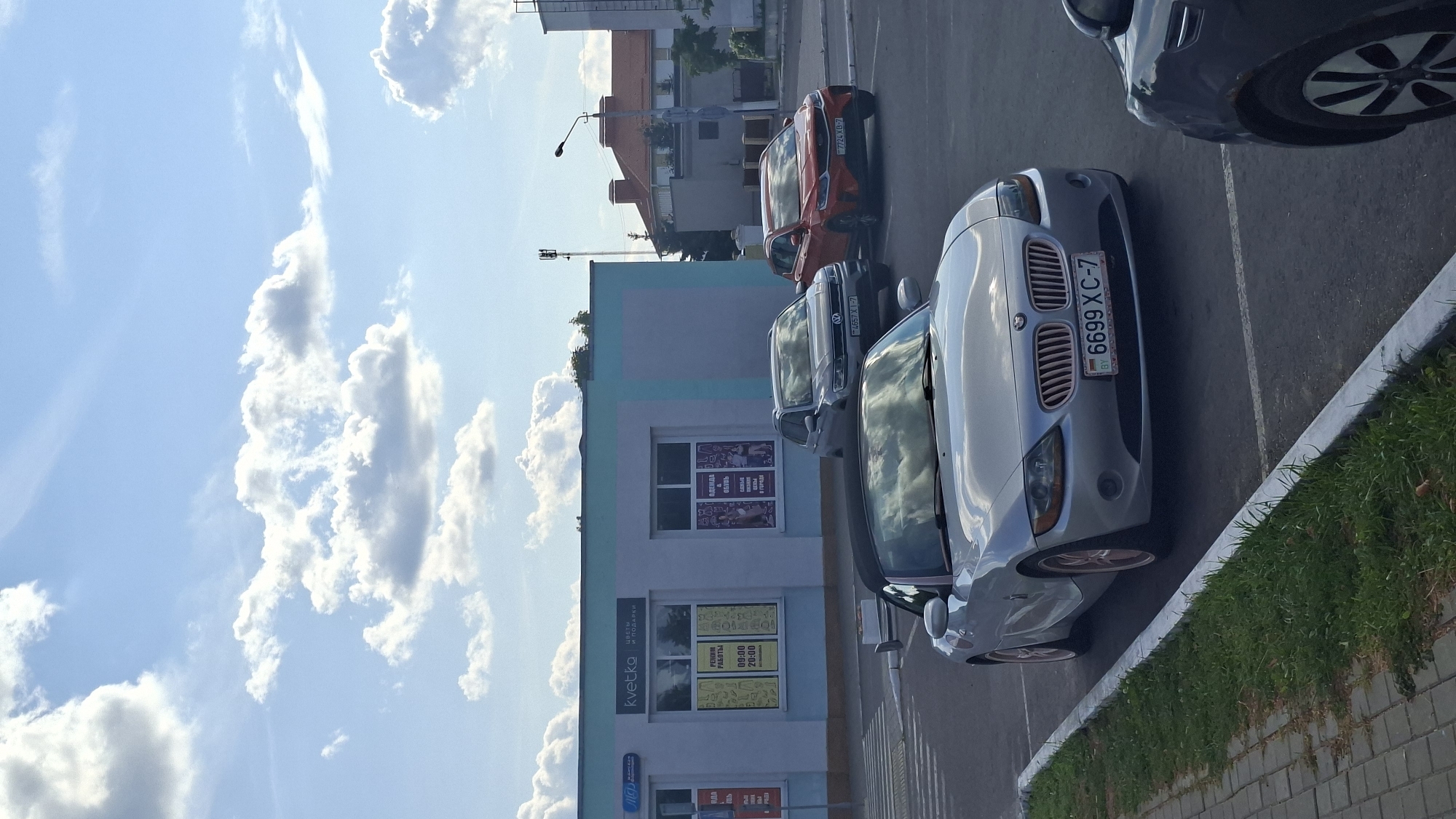 In Myadzyel, Belarus, on a clear day with scattered white clouds in a blue sky, a silver convertible BMW Z4 is parked in a lot adjacent to a light blue and white building. The building has signage for "kvetka" (flowers and gifts) and displays information about working hours and clothing and shoe sales. Other vehicles are present, including a silver SUV and an orange hatchback. A dark-colored car is partially visible in the foreground on the right. The foreground includes a paved walkway and a strip of grass.