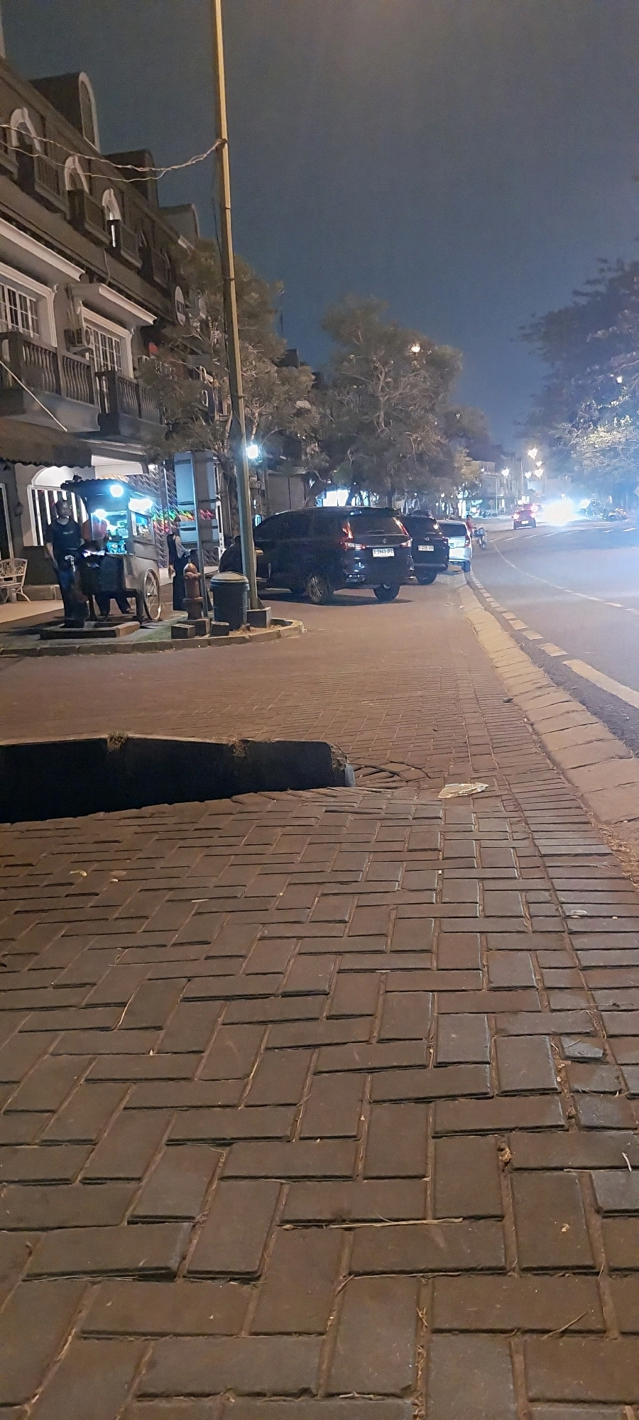 A low-angle, slightly blurry vertical shot captures a street scene at night in Kelapa Dua, Indonesia. The foreground is dominated by a paved walkway constructed of dark, rectangular bricks laid in a herringbone pattern. A dark, raised curb runs along the right edge of the walkway, separating it from the street. The street itself is visible with streetlights illuminating the scene, and traffic is implied by the headlights of distant vehicles.
To the left of the walkway, a building with a dark facade and multiple windows is partially visible. Above the ground level, balconies with white railings are present. A streetlamp on a pole stands between the building and the street. Next to the building, a food cart is set up, illuminated by bright lights, with at least two individuals standing near it, possibly vendors or customers. A fire hydrant and a trash bin are also visible on the walkway near the food cart.
Parked along the curb on the street are several vehicles, including at least two dark-colored vans. One van has a license plate that reads "B 3949 SP." The overall atmosphere is one of a typical urban night, with a mix of commercial activity and street infrastructure.