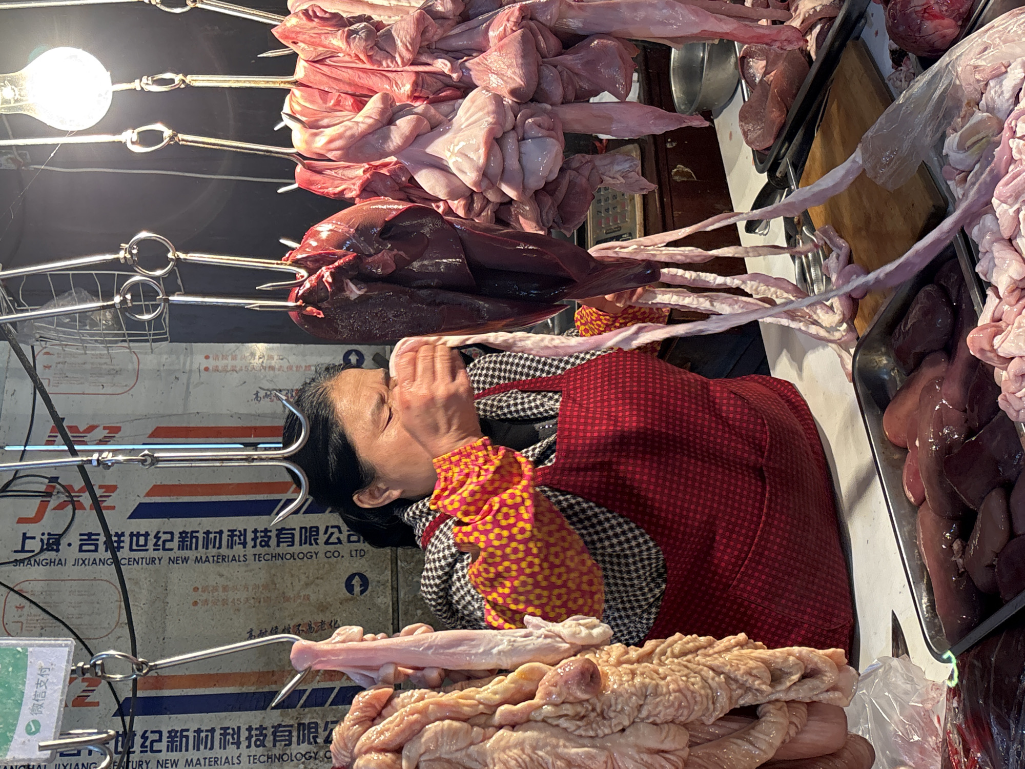 Here's a detailed description of the image:
**Scene:** An indoor market stall, likely in Jiangjin, China. The scene is dominated by raw meats and organs hanging from metal hooks. A woman wearing a red vest and patterned shirt is positioned in the center of the frame.
**Subjects:**
* **Raw Meats:** Various cuts and organs (potentially intestines, liver, lungs) are prominently displayed.
* **Woman:** An adult female, presumably a vendor, is positioned behind the meat display. Her hands are covering her face.
**Objects:**
* **Metal Hooks:** Used to hang the meat cuts for display.
* **Lightbulb:** A bare lightbulb illuminates the scene from above.
* **Printed Background:** A background with Chinese text and company information is visible. Likely advertising or signage for "Shanghai Jixiang Century New Materials Technology Co. Ltd."
* **Trays:** A metal tray filled with organs is visible on the right.
**Actions:**
* The woman has her hands covering her face.
**Environment:** The stall appears to be enclosed, likely part of a larger market space.