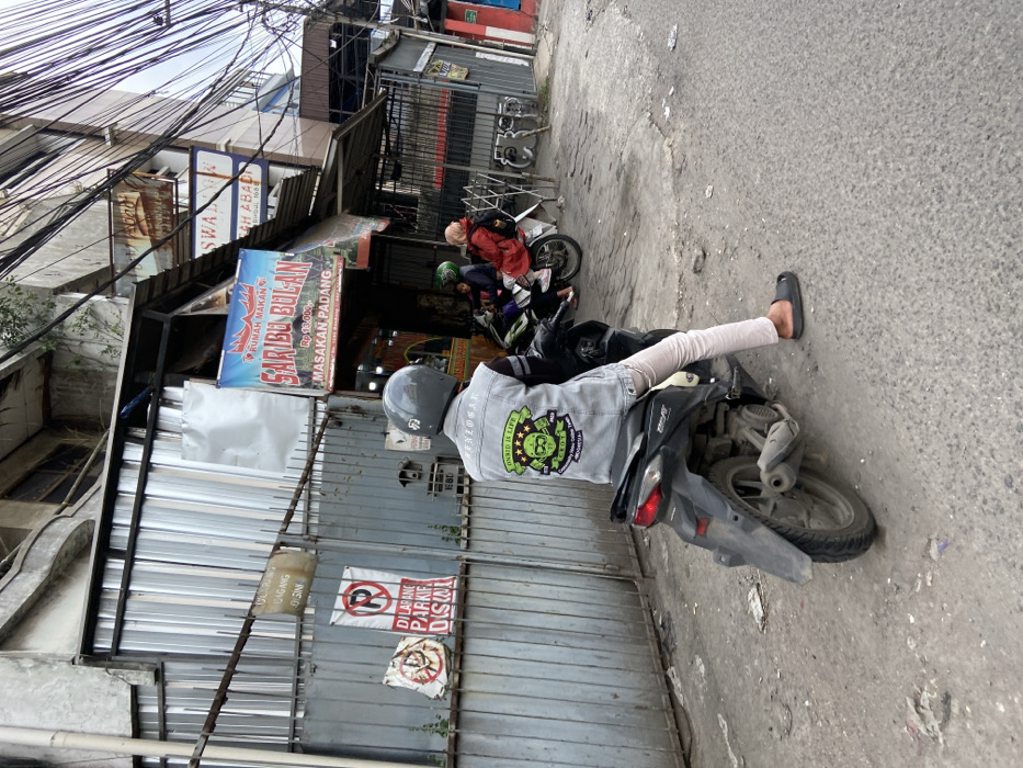 A man wearing a motorcycle helmet, a grey denim jacket with a green patch that says "Zombie Is Life," and beige pants sits on a black motorcycle. He is facing the left and looking down. The motorcycle is facing the right. The man is standing on the ground with his right foot on the motorcycle's foot peg and his left foot on the ground. The man is in front of a building with a sign that reads "Rumah Makan Saribu Bola" in red and white letters. The building is located at -6.965, 107.577 in Margahayu, Indonesia. There is another motorcycle to the right of the man, partially obscured by a car. There is a sign on the building to the left that reads "Dilarang Parkir Disini" in black and white. There are also some power lines strung overhead. The street is paved and dirty. There is a concrete curb on the right side of the road. The sky is overcast and the air is hazy. The man is parked in front of a restaurant.