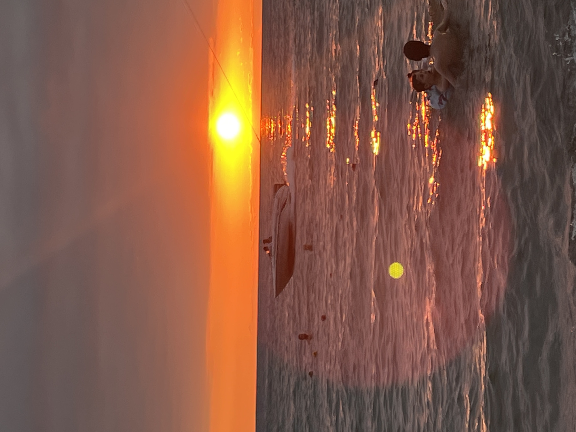 The image depicts a sunset over the Black Sea in Ureki, Georgia. A bright orange sun is setting on the horizon, casting a warm glow on the water and sky. Several people are visible in the water, some swimming and others standing. A speedboat is moving across the water, leaving a wake behind it. The water is calm, with gentle waves. There is a yellow-green circular object floating in the water. The overall atmosphere is serene and peaceful.