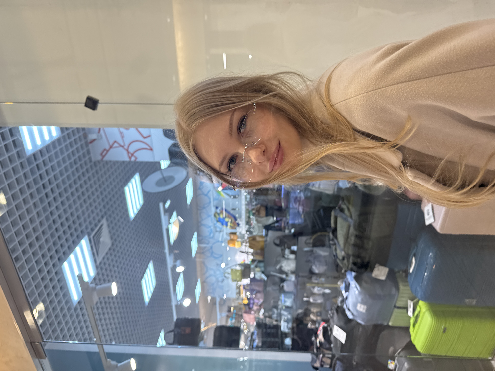 A young, blond-haired woman with glasses is reflected in a glass window, which is likely a storefront. The reflection shows the interior of what appears to be a store selling luggage and bags, with various items displayed on shelves. Through the window, part of the ceiling is visible, showing a grid pattern with integrated lighting fixtures. This image was taken in Perm, Russia.