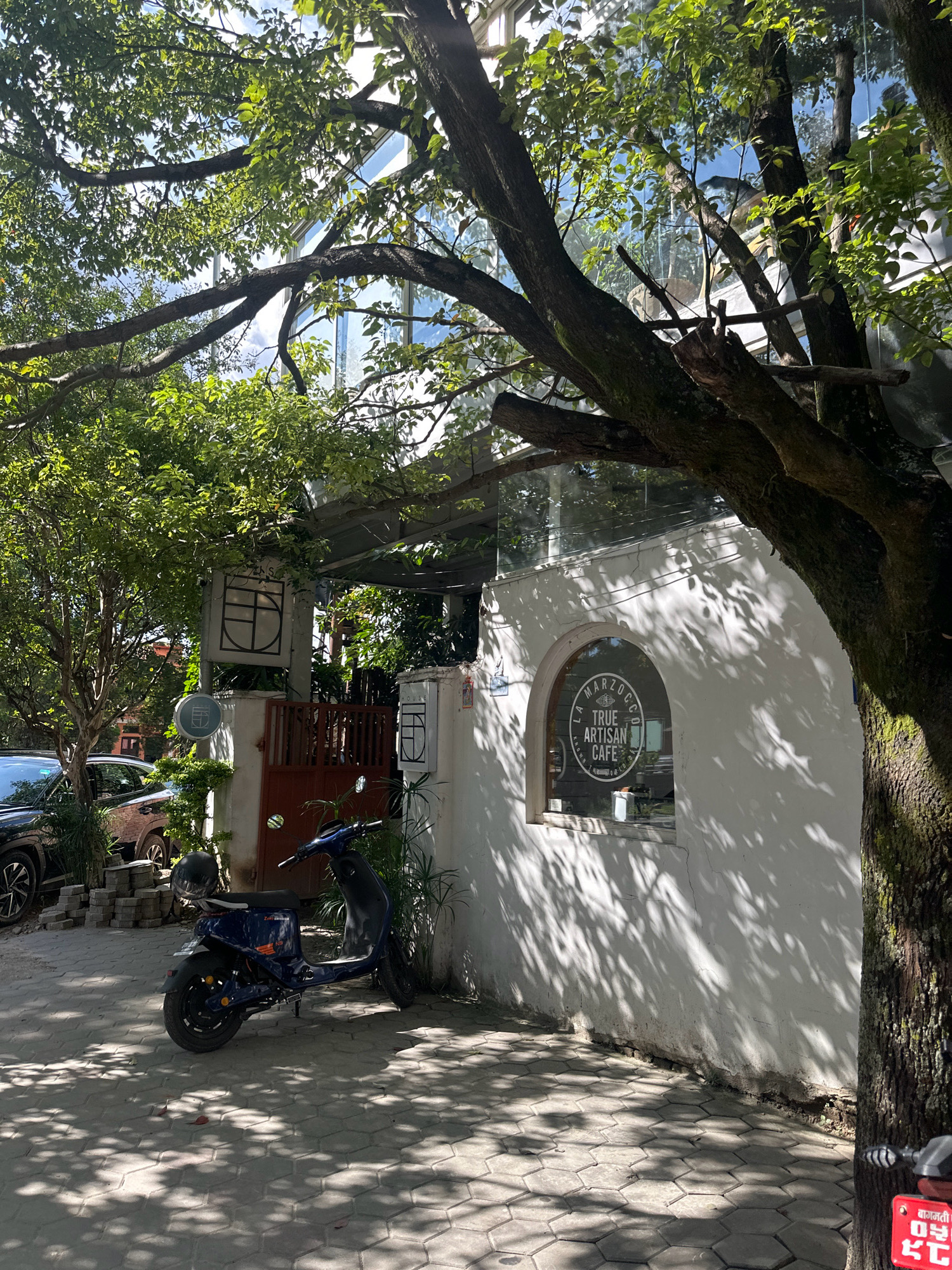 The image depicts a street scene in Lalitpur, Nepal, under dappled sunlight filtering through trees. In the foreground, a blue electric scooter with a black helmet on its seat is parked on a hexagonal paved pathway. To the right of the scooter, a white building with an arched window is visible. The window has a circular logo that reads "LA MARZOCCO TRUE ARTISAN CAFE". To the left of the scooter, a dark-colored car is partially visible, along with a stack of bricks. Behind the car and scooter, a brown gate is set into a wall, and further back, a modern building with large glass windows can be seen through the foliage of trees. The overall scene is a blend of urban elements and natural greenery, characteristic of a cafe or commercial area in Lalitpur.