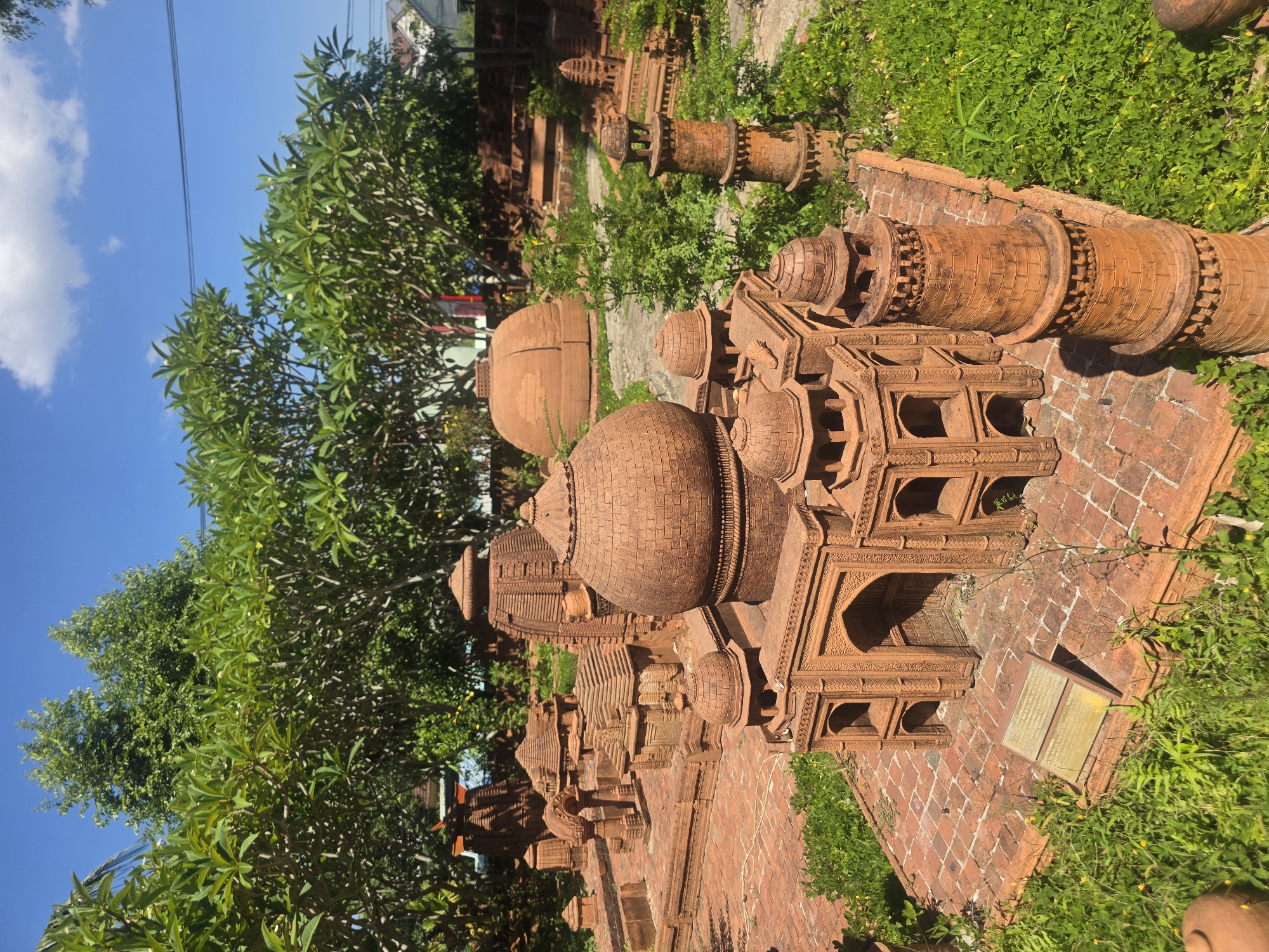 This image captures an outdoor scene showcasing an extensive collection of miniature terracotta architectural models, resembling ancient temples or religious structures. The models are intricately detailed, featuring domes, multi-tiered roofs, arched openings, and decorative patterns, suggesting styles reminiscent of various South and Southeast Asian architectures. They are arranged on a brick-paved ground, interspersed with vibrant green grass and small yellow flowers.
Lush green trees, including some identifiable as frangipani (plumeria) with their distinct leaf shape, frame the display, providing natural shade. The sky is a clear blue with scattered white clouds, indicating a bright, sunny day, likely midday or early afternoon given the strong sunlight and shadows. Faint power lines are visible against the sky.
In the foreground, a rectangular plaque, likely providing information about the exhibit, is visible on the brick surface next to one of the more elaborate models; its text is unreadable. The scene presents a curated display of craftsmanship and cultural heritage in a peaceful, garden-like setting. The location context provided is Đà Nẵng, Vietnam.