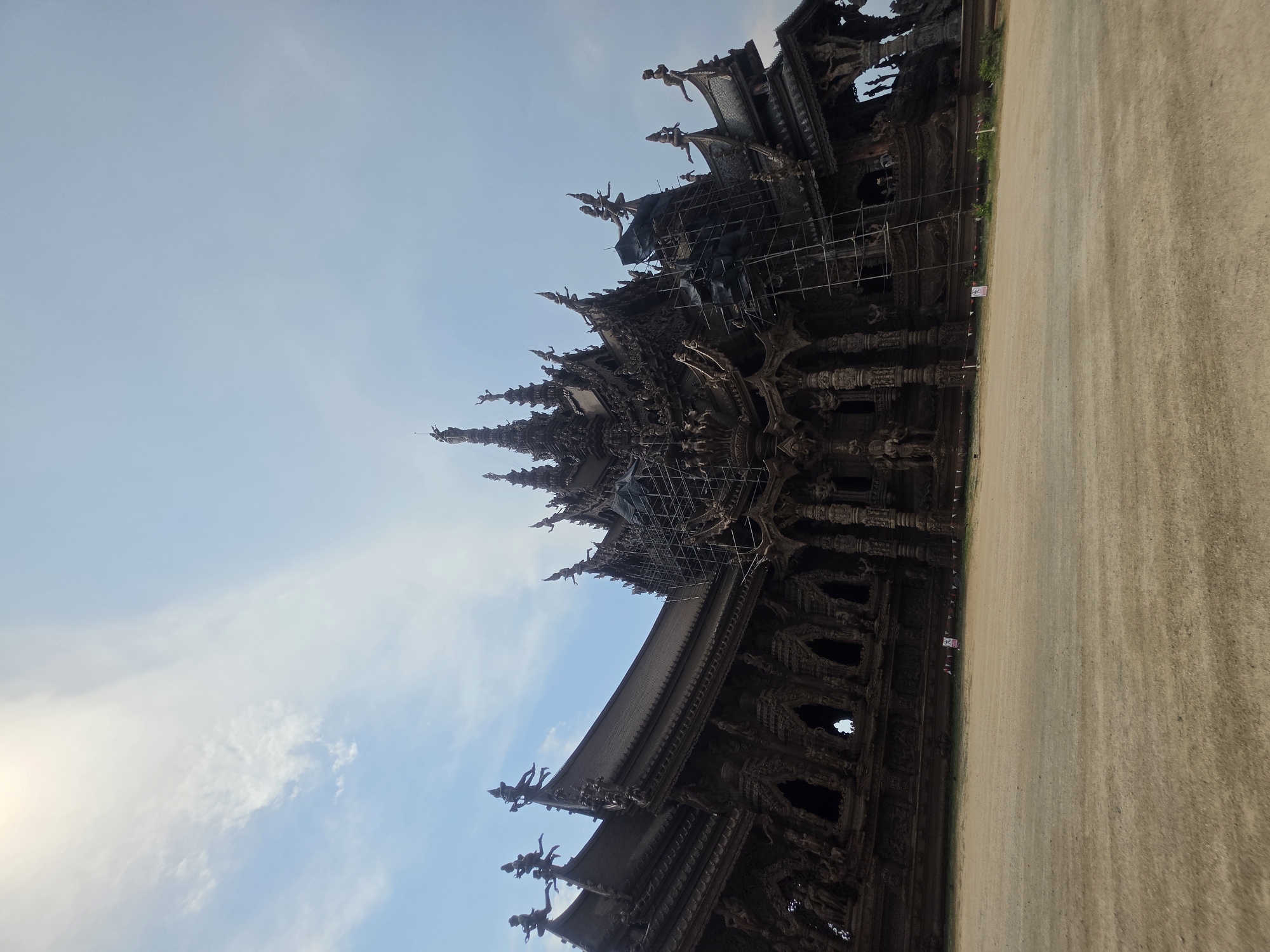 The image displays a majestic, large-scale structure constructed entirely of dark, intricately carved wood, set against a clear blue sky with scattered white clouds. The building, consistent with the architectural style found in Pattaya City, Thailand, features multiple spires, ornate rooflines, and countless detailed sculptures. These carvings depict a variety of figures, including mythical creatures, deities, and human-like forms, adorning nearly every surface of the structure, from its columns to its eaves.
Parts of the building are currently undergoing renovation or construction, indicated by extensive scaffolding wrapped around sections of the upper levels and spires. This suggests an ongoing effort to maintain or expand the elaborate wooden edifice. The general lighting suggests it is daytime, likely late morning or early afternoon, with ample natural light illuminating the scene.
In front of the structure, a wide, flat expanse of light brown, sandy ground stretches across the lower and right portions of the image. A narrow strip of green vegetation, possibly small bushes or plants, runs along the base of the wooden building, separating it from the sandy area. A small, rectangular white sign with red markings is visible on the sandy ground near the base of the structure, but its specific text or symbols are too indistinct to read clearly. No people are discernible in the frame, highlighting the grandeur of the architecture against the open sky and ground.