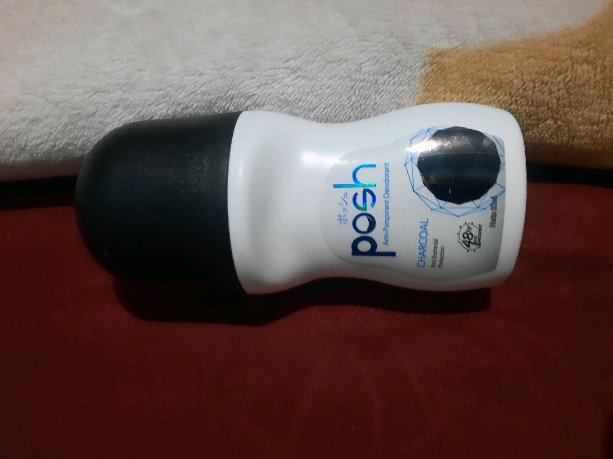 A white and black roll-on deodorant is positioned horizontally on a red fabric surface, with a beige towel draped behind it. The deodorant bottle is predominantly white with a black cap. The label on the bottle features the brand name "posh" in a gradient blue font, with Japanese characters above it. Below "posh", it reads "Anti-Perspirant Deodorant". Further down the label, "CHARCOAL" is written in uppercase, followed by "Anti Bacterial Protection". A small graphic indicating "48hr" protection is also visible. The scene appears to be an indoor setting, likely a home, given the presence of a towel and fabric surfaces. No people or specific landmarks of East Jakarta are identifiable in the image.
