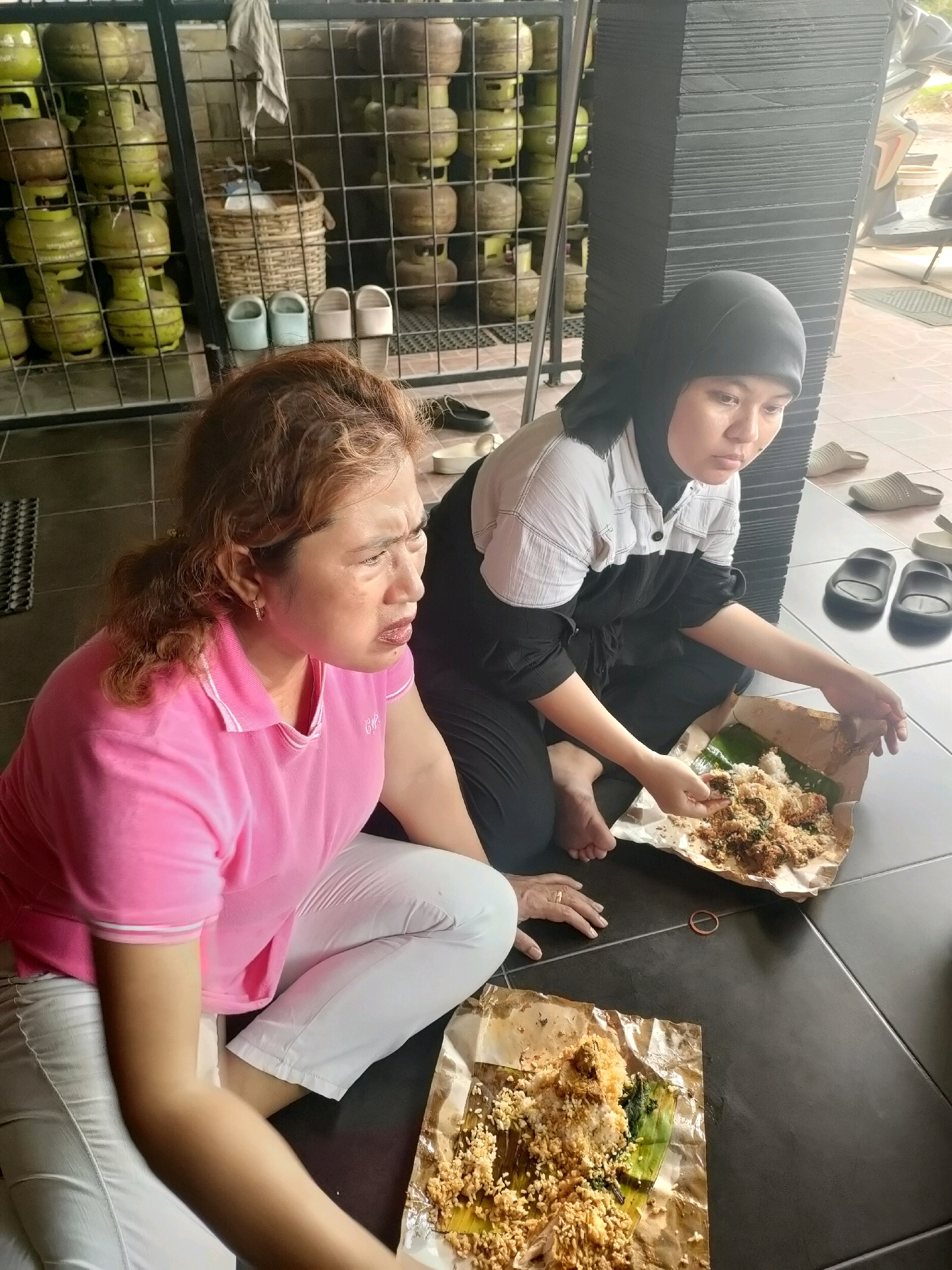 Two Indonesian women are sitting on a tiled floor, eating from paper-wrapped meals. The woman on the left, wearing a pink polo shirt and white pants, has reddish-brown curly hair and is looking to her right with a concerned expression. The woman on the right wears a black headscarf and a black and white shirt with black pants. She is actively eating from her meal. In the background, numerous green and brown gas cylinders are stacked in metal shelving units, suggesting a storage or retail area. The overall environment appears to be an outdoor or semi-outdoor space with a tiled floor and a dark-colored textured wall. Based on the visual cues of the gas cylinders commonly used in Indonesia and the demographic of the individuals, the scene is likely located in Pekanbaru, Indonesia.