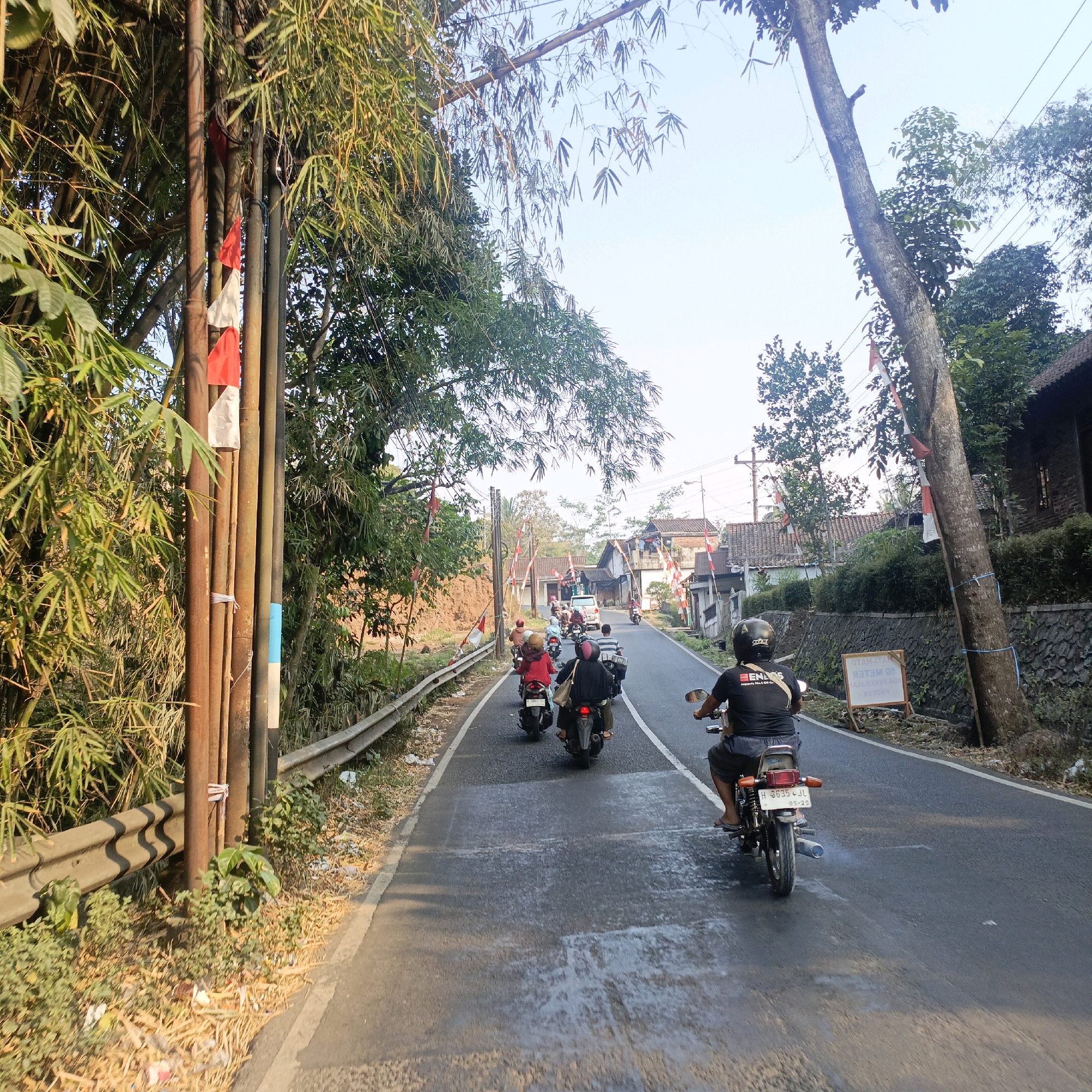 The image depicts a street scene in Salatiga, Indonesia, likely in a rural or suburban area given the presence of bamboo groves, trees, and houses with traditional tiled roofs. A group of people are riding motorcycles in a convoy down a paved road. The road has a white line marking the center and a guardrail on the left side. Several Indonesian flags, red and white, are visible, attached to poles or trees along the roadside, suggesting a national celebration or commemoration. The sky is clear and the sunlight indicates it is daytime, possibly late afternoon due to the long shadows. The motorcycles are carrying multiple passengers, some wearing helmets. The environment is lush with green foliage and some dry grass and debris on the roadside. There are several houses visible in the background and a white sign with text written in Indonesian.