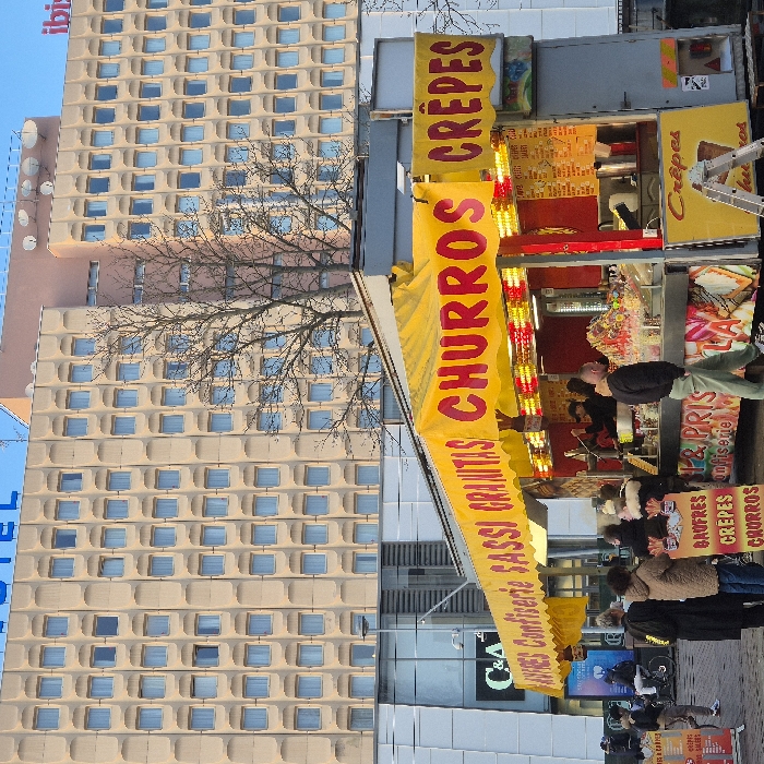 The image depicts an outdoor urban scene featuring a multi-story building and a vibrant food stall, likely at dusk or early evening, given the illuminated stall and the visible sky.
To the left, a tall, beige or light brown building with numerous square windows, each framed by a textured recess, dominates the background. The "ibis" hotel logo is visible at the very top left, and the word "HOTEL" is partially visible in a vertical format near the bottom left edge of the building. In front of this building, a large deciduous tree with bare branches suggests cooler weather, possibly autumn or winter.
The focal point of the image is a brightly lit food stall positioned in the foreground to the right. The stall is adorned with yellow awnings and banners featuring large red text. The main banner clearly displays "CHURROS" and "CREPES". A lower banner further reads "AVES Confiserie SASSI GRANITAS CHURROS CREPES". Additional smaller signs on the stall's structure advertise "CREPES," "BAUFRES," "CHURROS," "Confiserie," "GRANITAS," and "& PRIS." The interior of the stall is brightly illuminated with numerous small lights, showcasing various sweets, candies, and potentially ingredients for the advertised items. A small ladder is leaning against the right side of the stall.
Several people are gathered in front of the stall, appearing to be customers. They are dressed in coats, reinforcing the impression of cool weather. One person is visible behind the counter, likely a vendor. The activity suggests ordering or waiting for food.
In the background, behind the food stall and below the large hotel building, another building with a "C&A" logo is partially visible. The ground is paved, indicating a street or market setting. Near the bottom right, a blue advertising board and a bicycle can be seen, further contributing to the urban environment. The overall atmosphere is lively due to the bright lights and presence of people.
