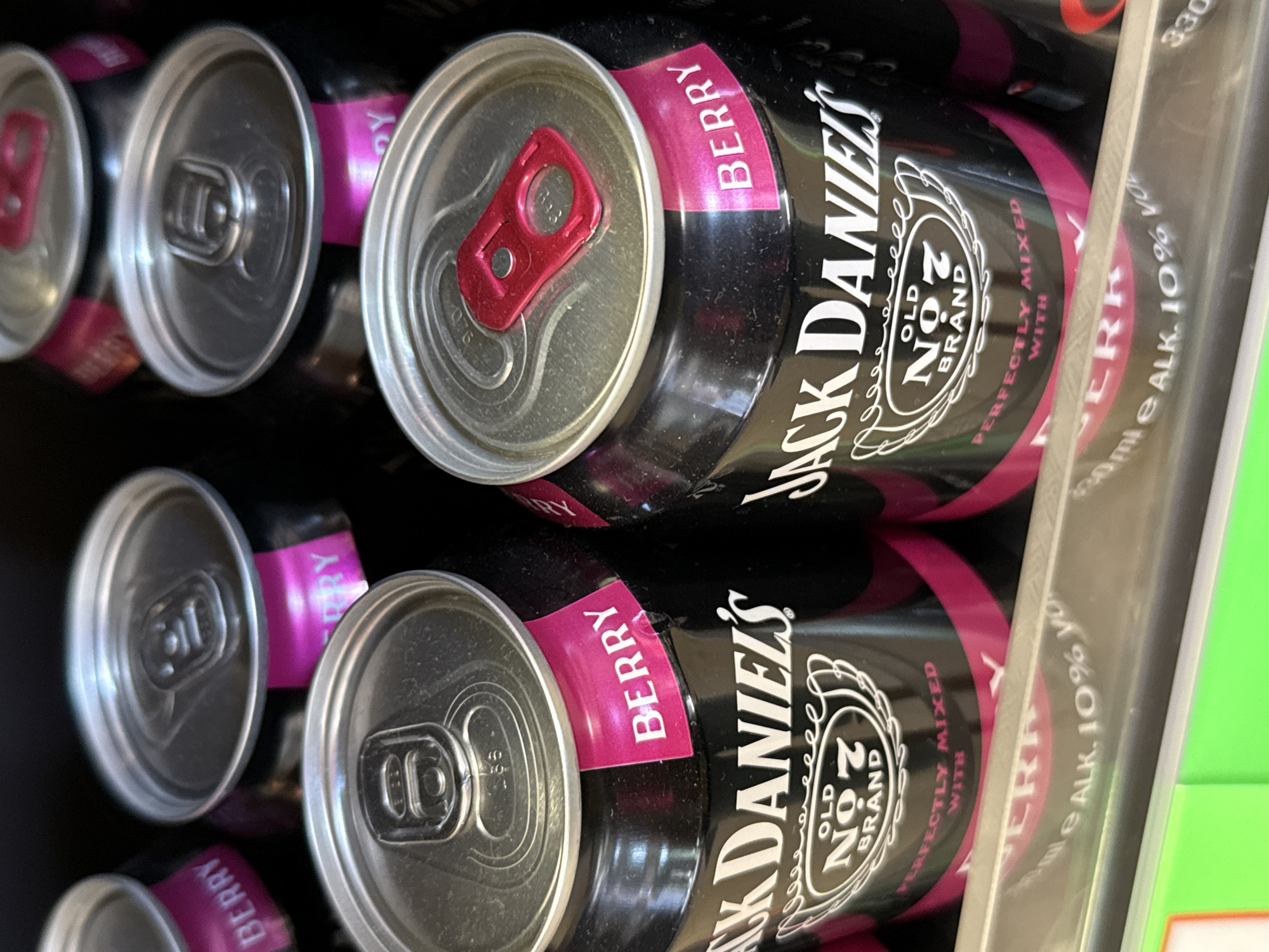 The image shows a close-up of several cans of Jack Daniel's Old No. 7 Brand Berry flavored beverage. The cans are black with white lettering and a pink band around the top with the word "BERRY" printed on it. The top of the cans are silver with red pull tabs. The cans are arranged in rows, suggesting they are on a shelf in a store. The background is dark, indicating a dimly lit environment. The bottom of the image shows a silver shelf with some text and a bright green surface below. The text on the shelf appears to be related to the beverage's volume and alcohol content, reading "330 ml e ALK. 10% vo". This scene is likely taking place in a supermarket or convenience store in Bad Lauterberg im Harz, Germany, given the context provided.